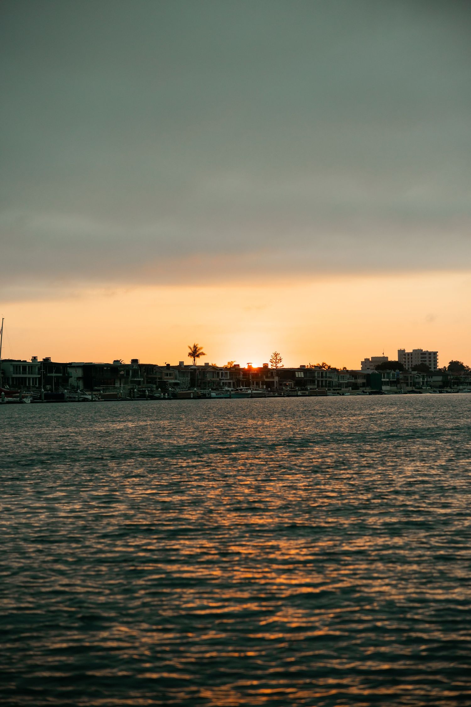 Dramatic sunset over calm ocean waters with silhouetted buildings along the coastal horizon.