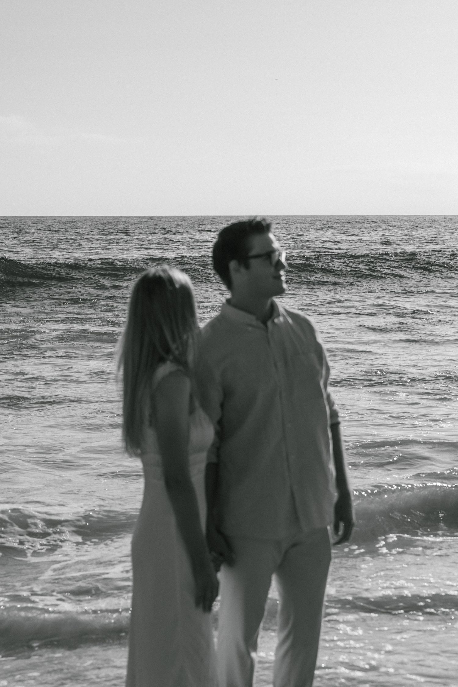 A black and white photo of a couple standing together on a beach with waves in the background.