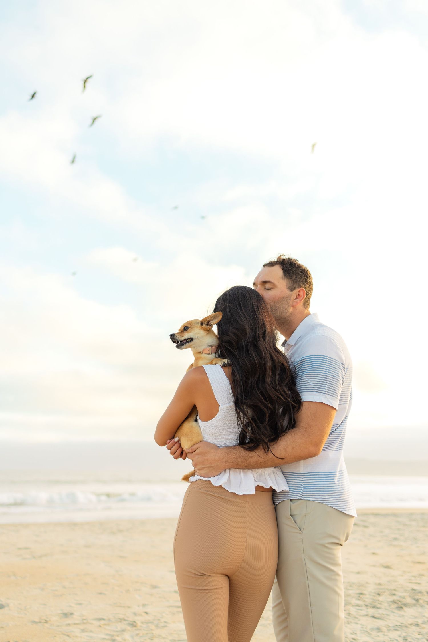 A couple embraces on a sunny beach while holding their small dog during a romantic photoshoot.