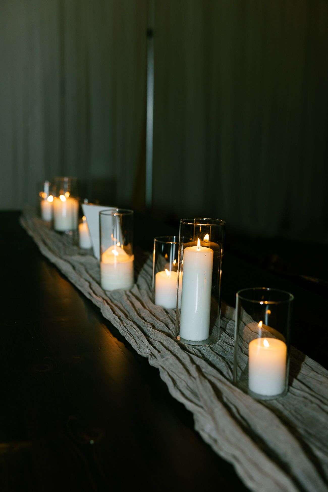 A row of glowing white candles in glass holders arranged on a rustic wooden plank.