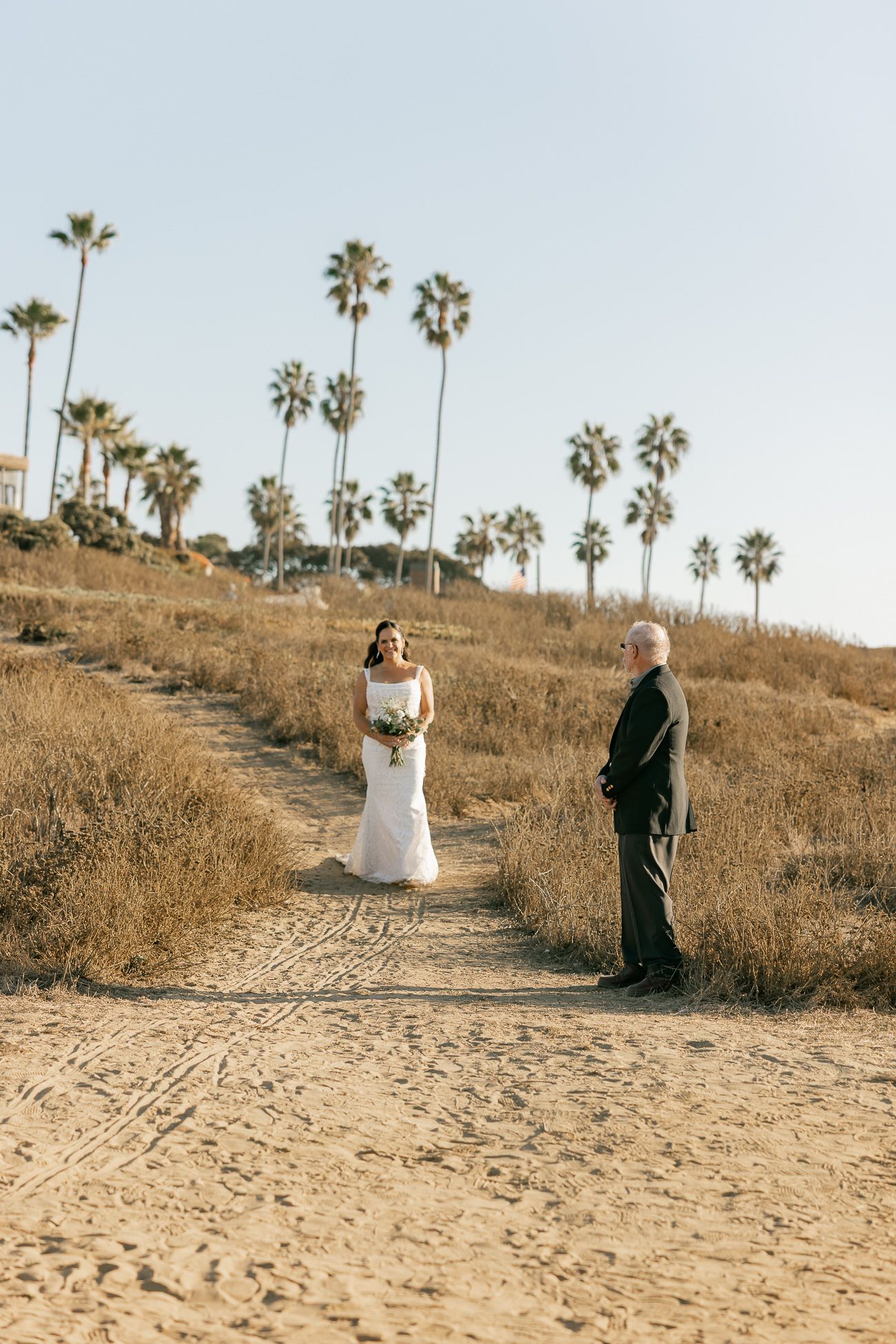 A couple stands on a sandy path with tall palm trees dotting a hillside during a romantic outdoor photoshoot.