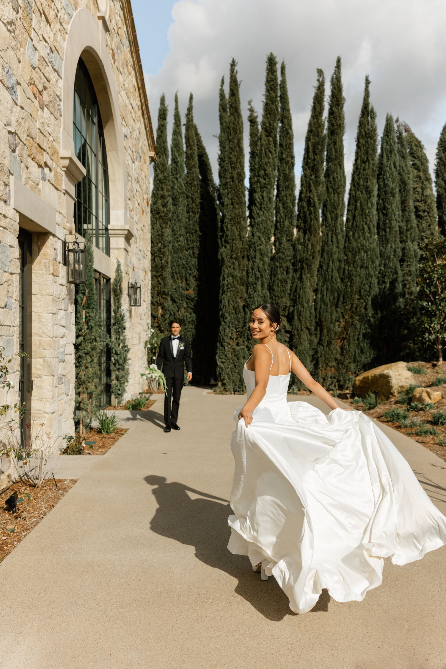 Wedding couple walking along stone building path lined with tall cypress trees.