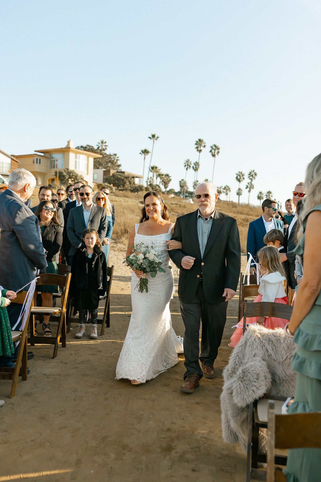 Outdoor wedding procession with couple walking down aisle surrounded by palm trees and guests on a sunny day.