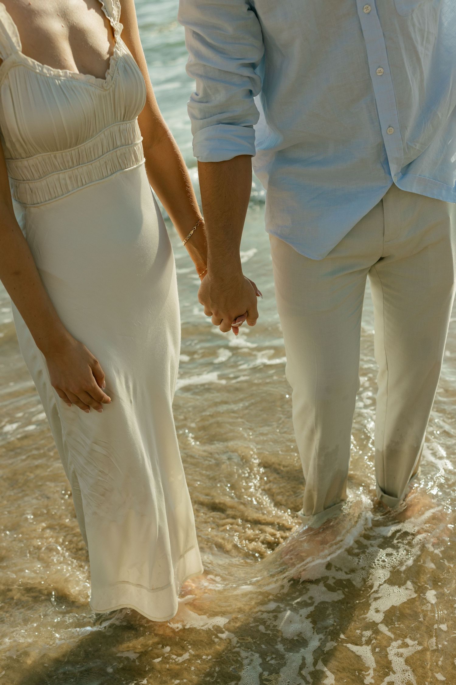 A close-up shot of a couple holding hands while standing in shallow ocean waves wearing white formal attire.