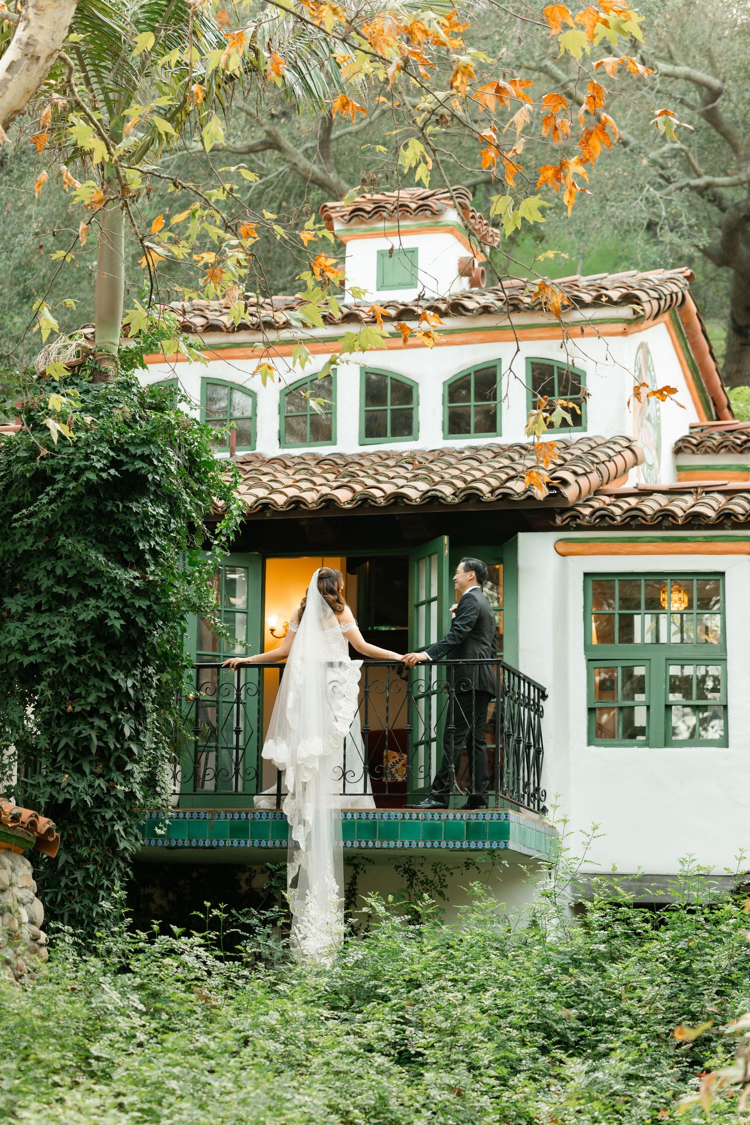 Charming Spanish-style house with terracotta roof tiles features a green balcony surrounded by lush foliage and orange flowers.
