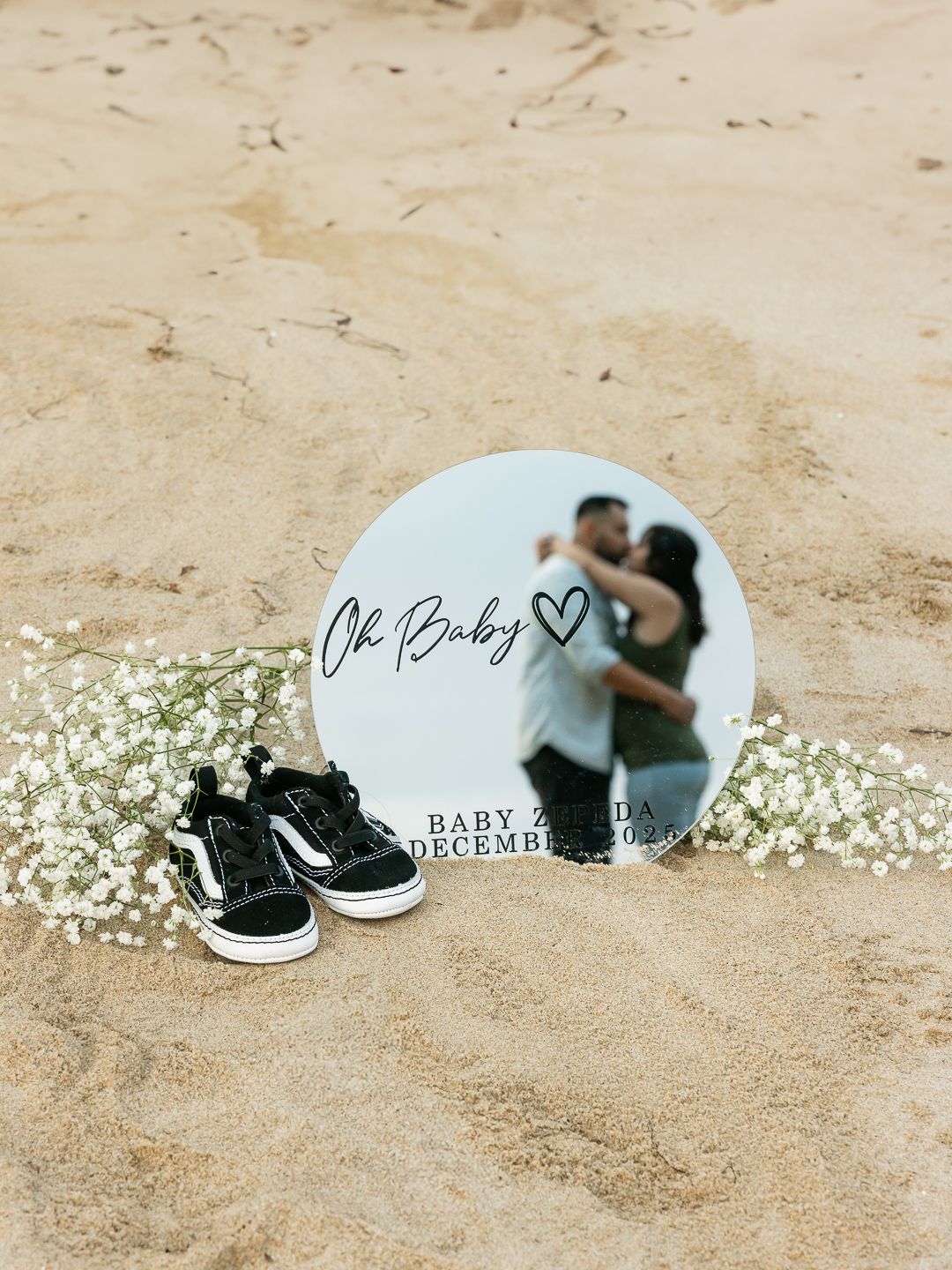 A baby announcement photo collage showing black sneakers and baby shoes with delicate white flowers on sandy ground.