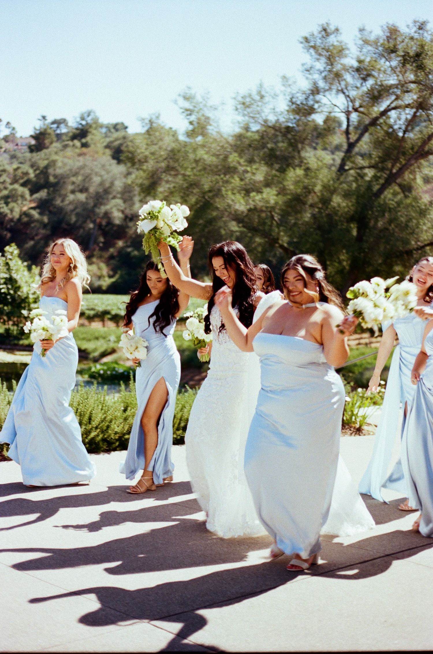 Bridesmaids in flowing white dresses pose together in a sunny outdoor setting during a wedding celebration.