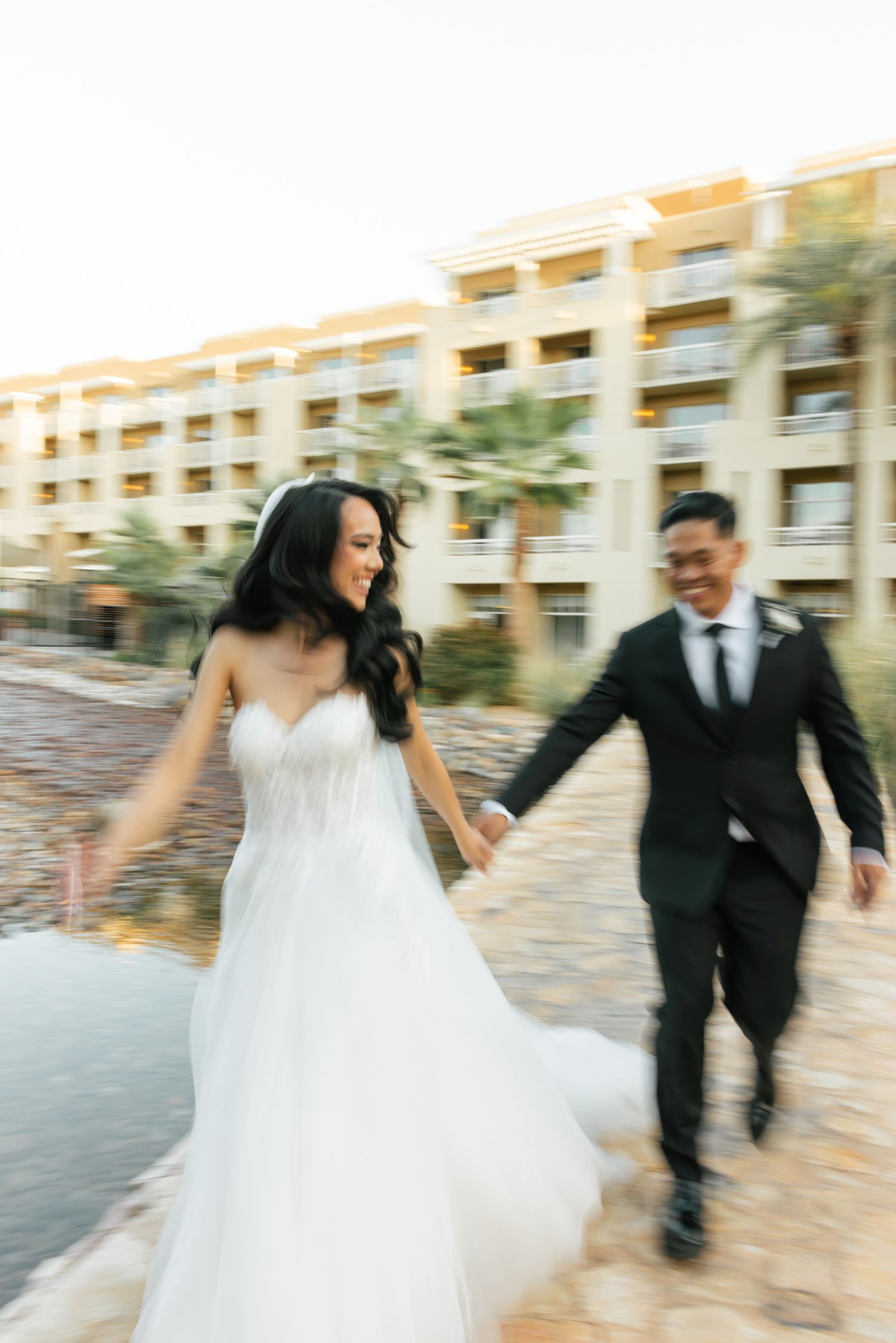 A couple in wedding attire runs joyfully along a stone pathway at a resort, creating a dynamic motion blur effect.