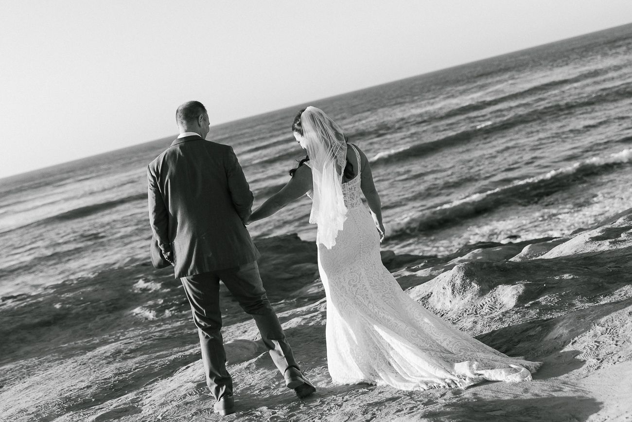 Black and white photo of a couple standing on rocky ocean shoreline with waves crashing.