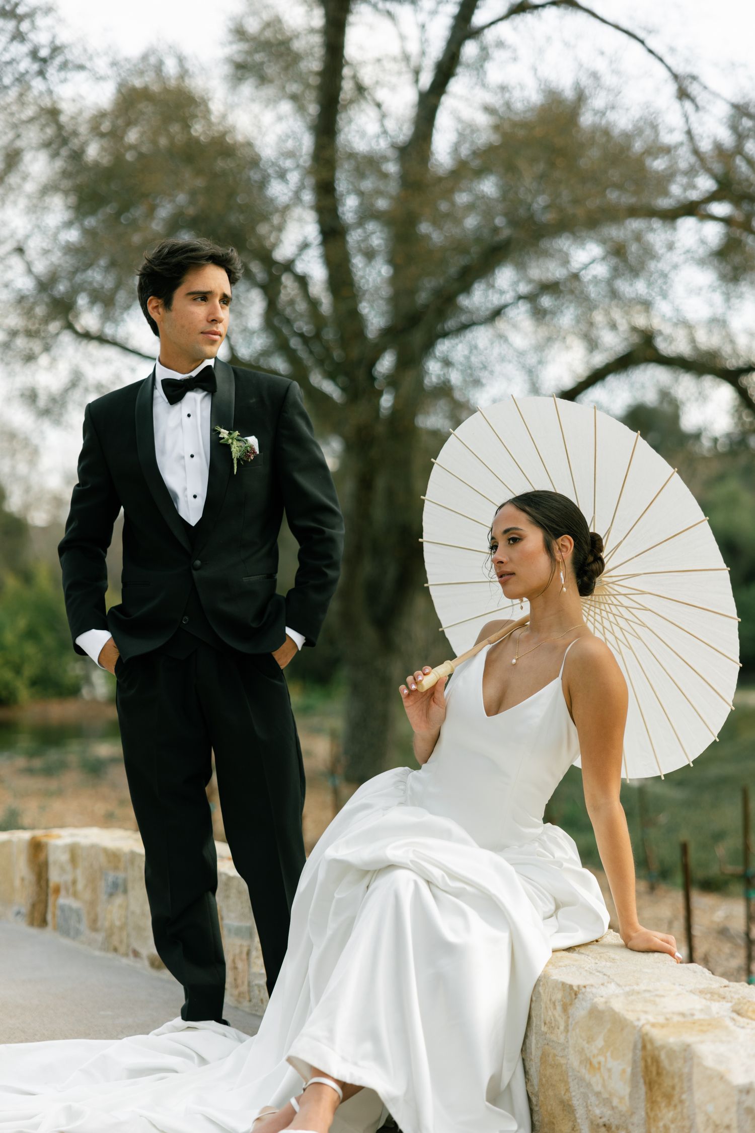 A wedding couple poses with a white parasol against a rustic stone wall backdrop.