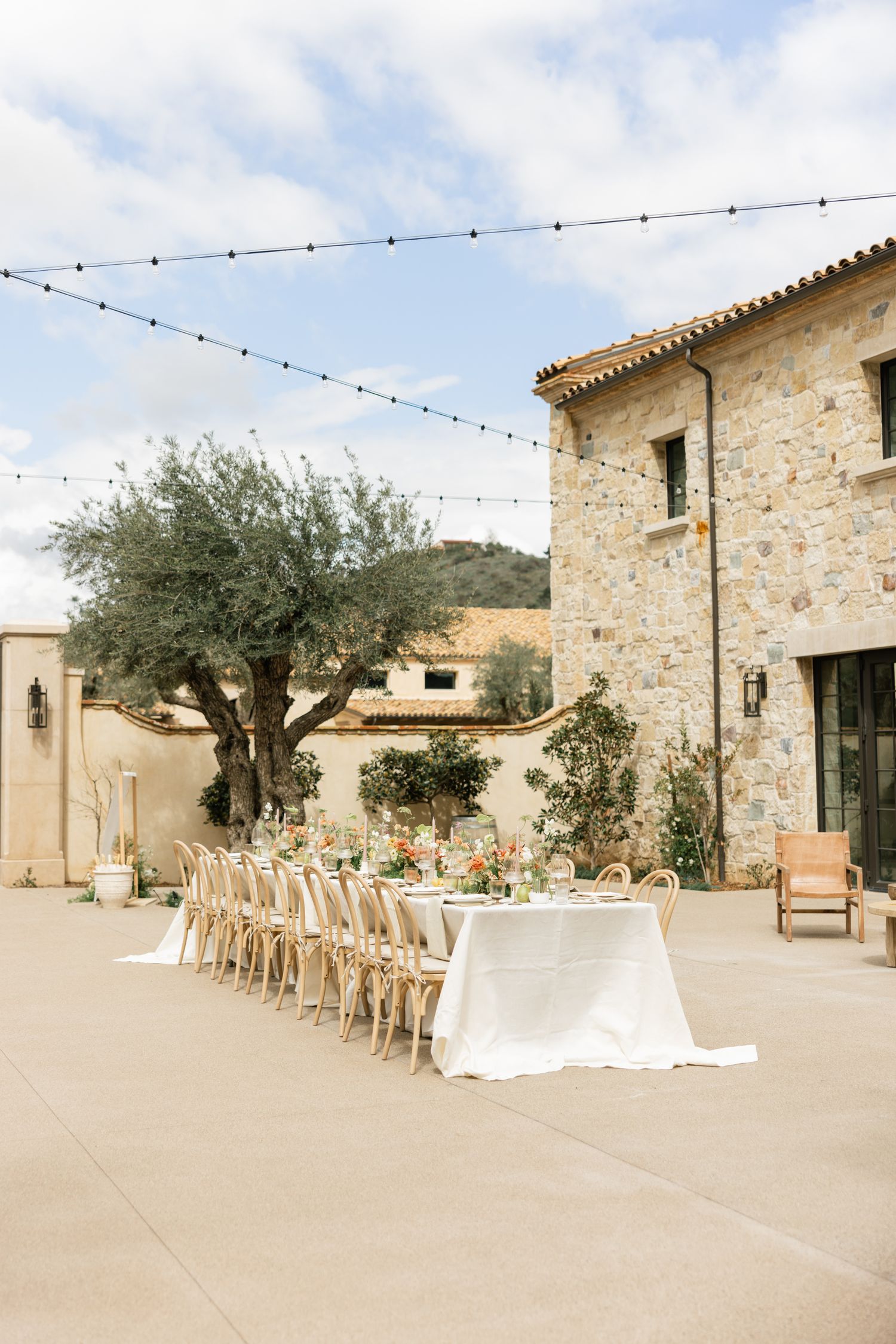 Outdoor wedding reception setup with long table and string lights at a rustic stone villa with olive trees.