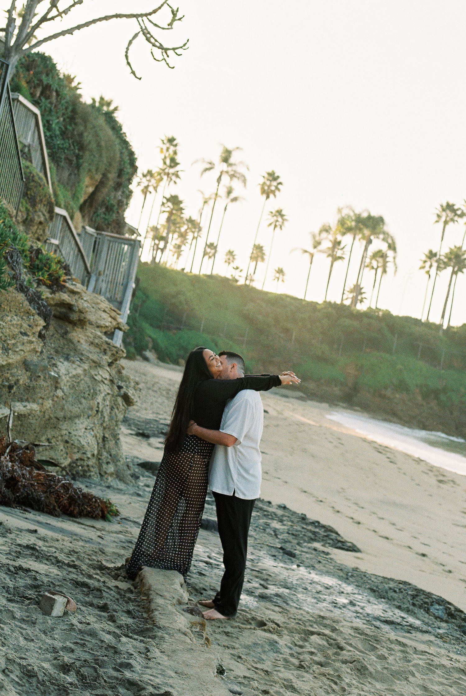 A romantic couple embraces on a scenic beach with palm trees and cliffs during golden hour.