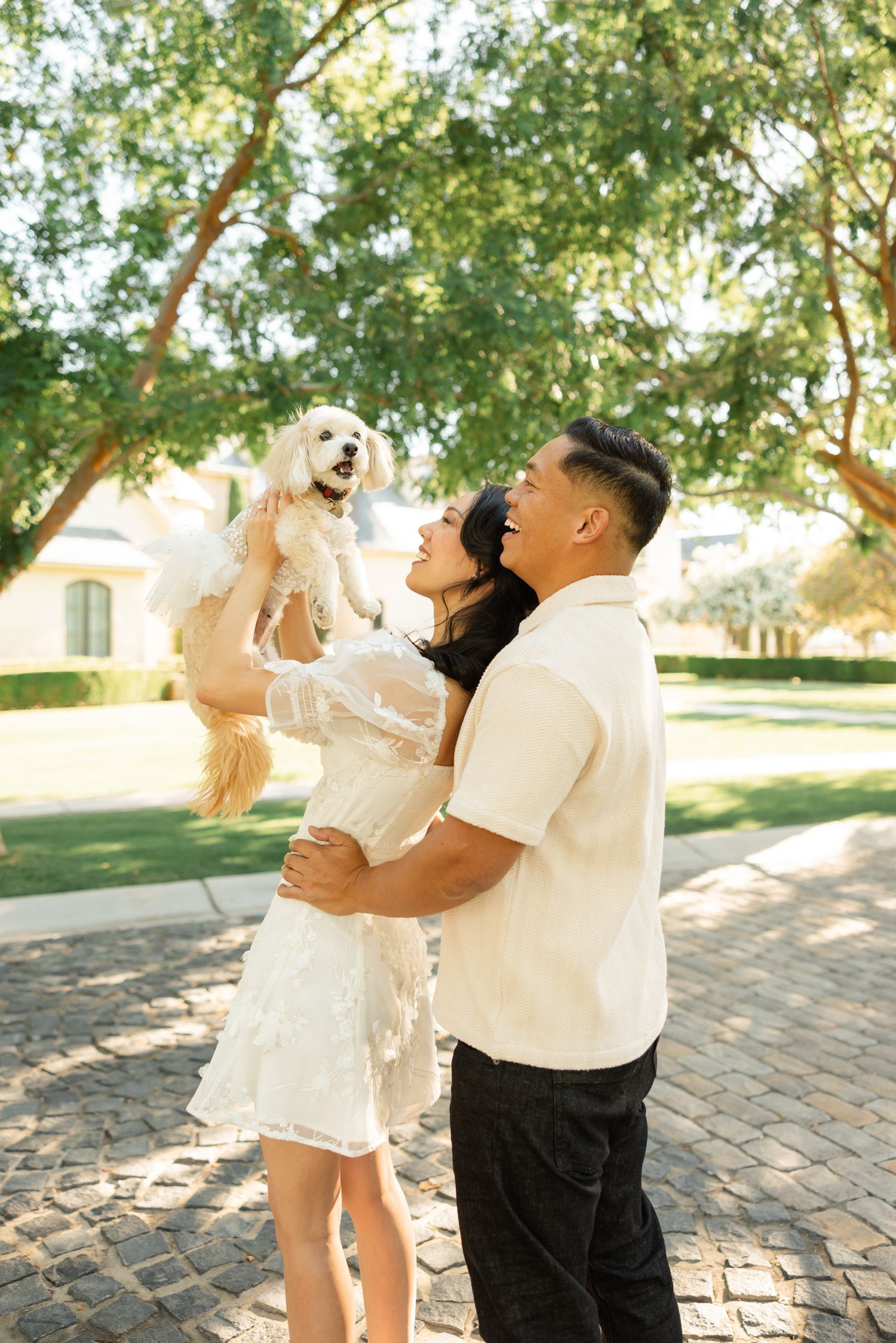 A couple shares a joyful moment while dancing together under green trees on a sunny day.