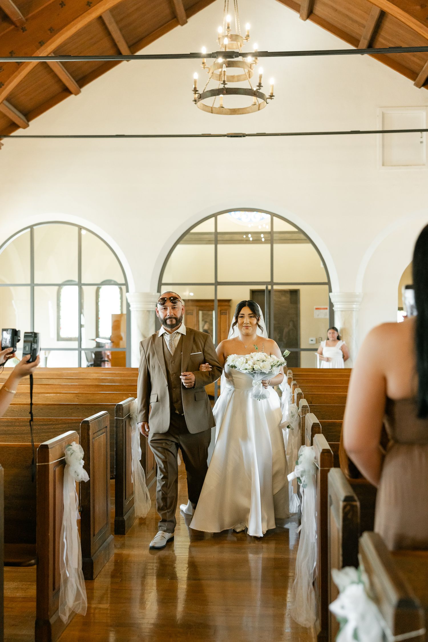 A wedding ceremony in a traditional church with wooden pews and arched windows features decorative white bows along the aisle.