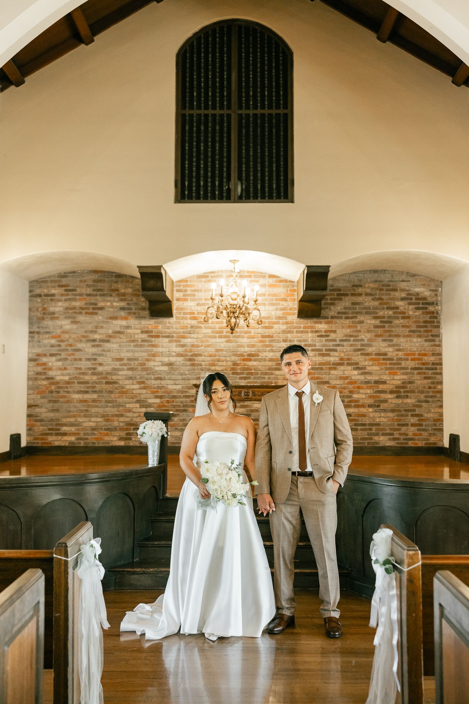 A traditional church interior with exposed brick walls and wooden pews is decorated for an intimate wedding ceremony.