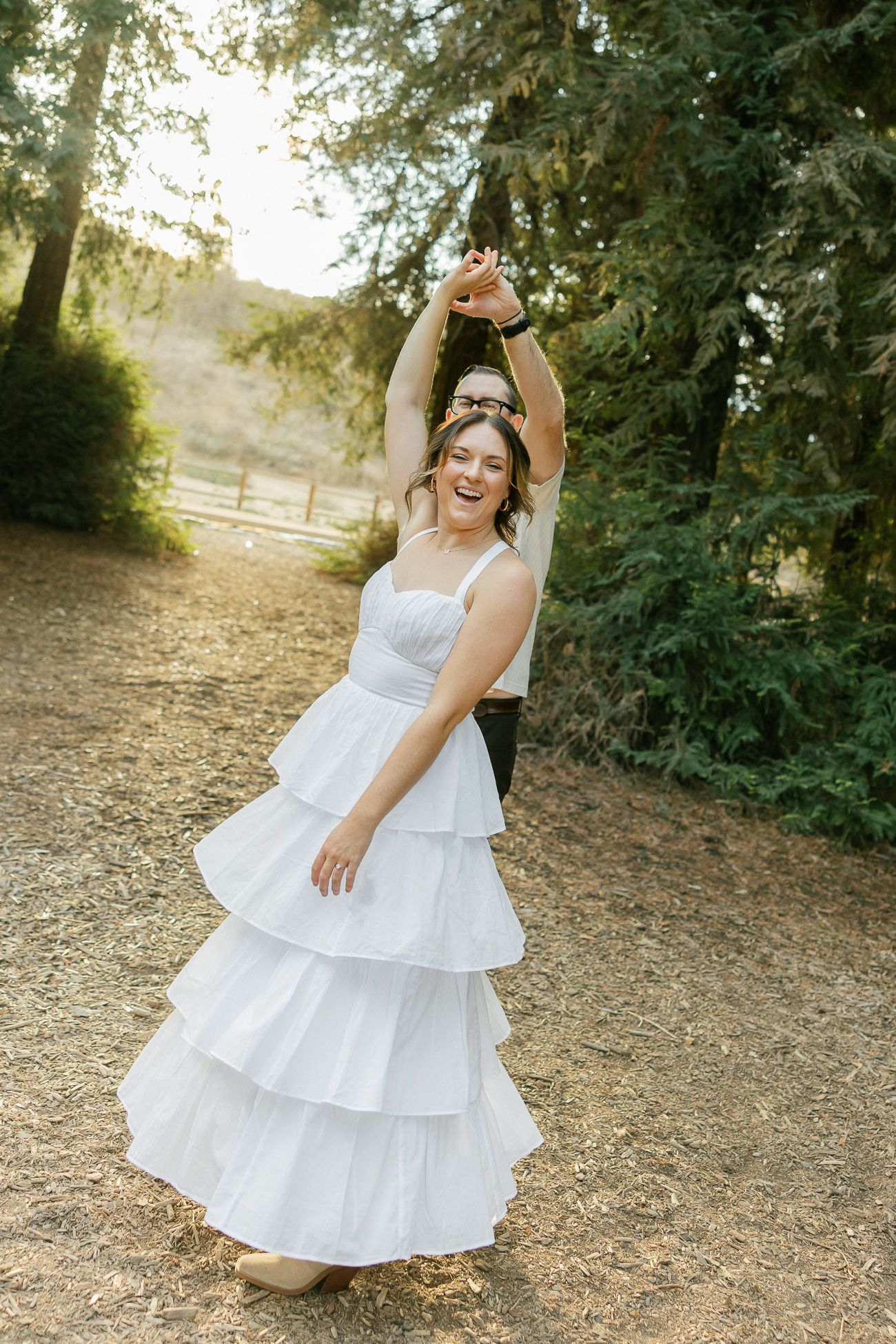 A bride in a tiered white wedding dress twirls joyfully on a forest path during golden hour.