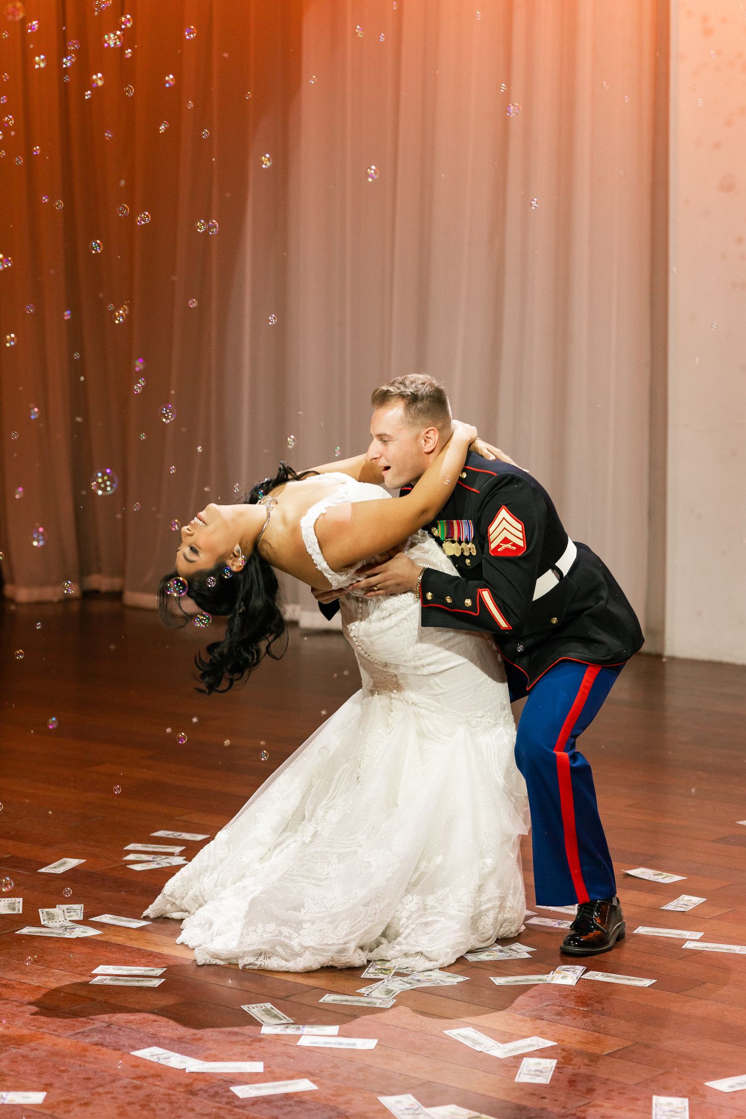 Couple in formal attire dancing dramatically on a wooden floor with falling confetti and a curtained backdrop.
