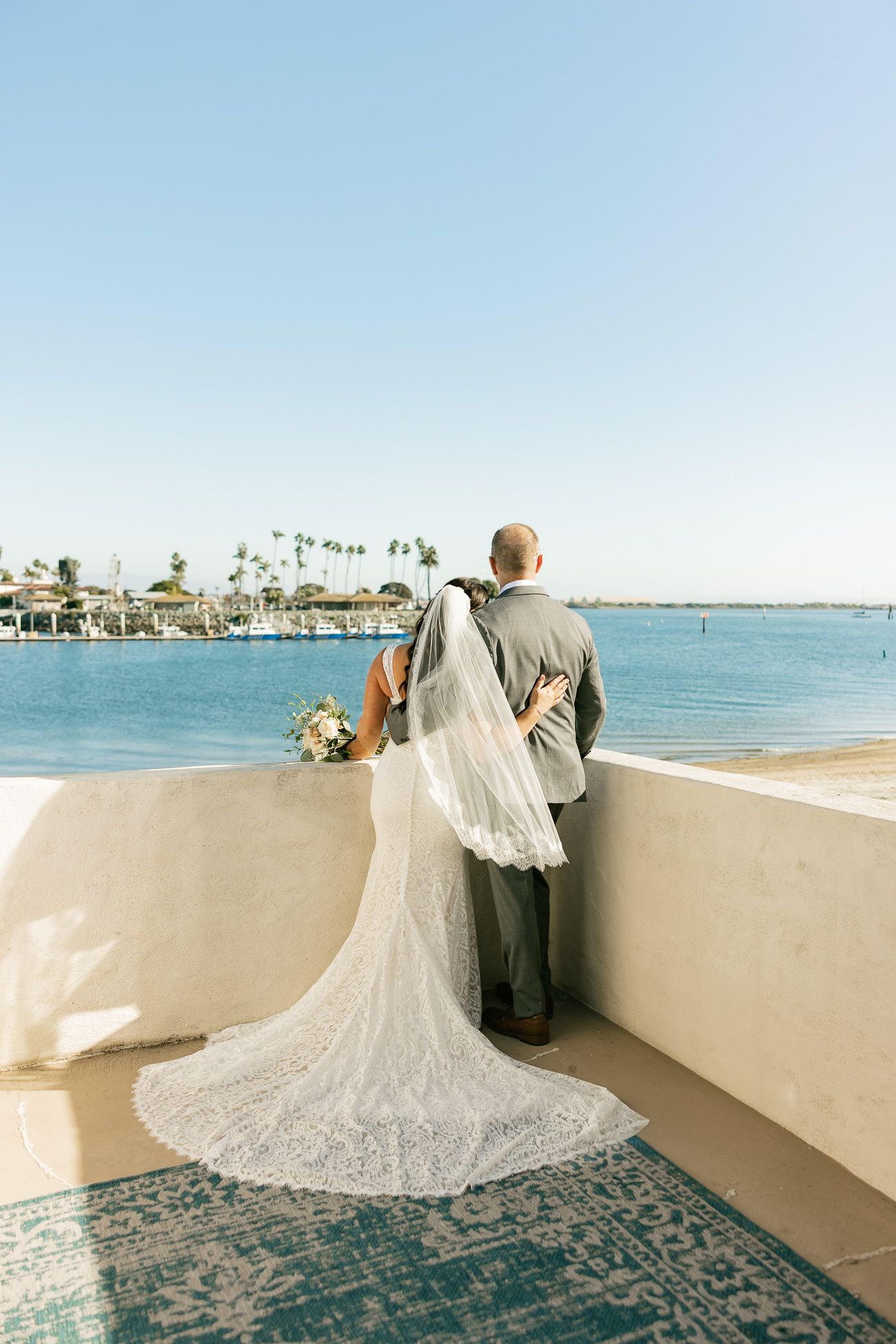 Couple shares romantic moment on seaside balcony with bride's lace train flowing behind on decorative rug.