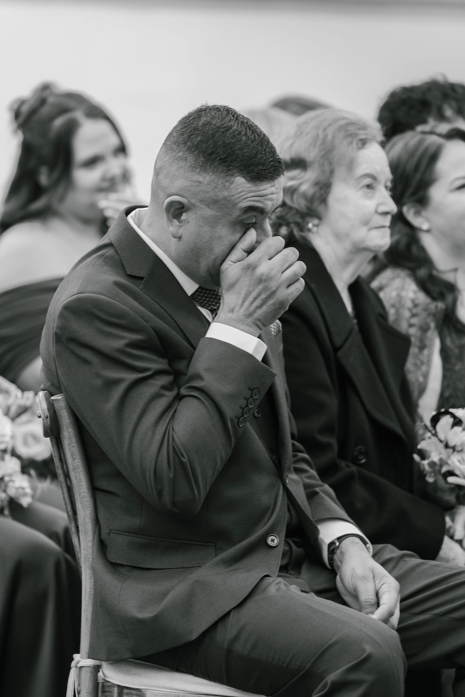 An emotional moment at a wedding ceremony shows a suited man wiping tears while seated among guests.