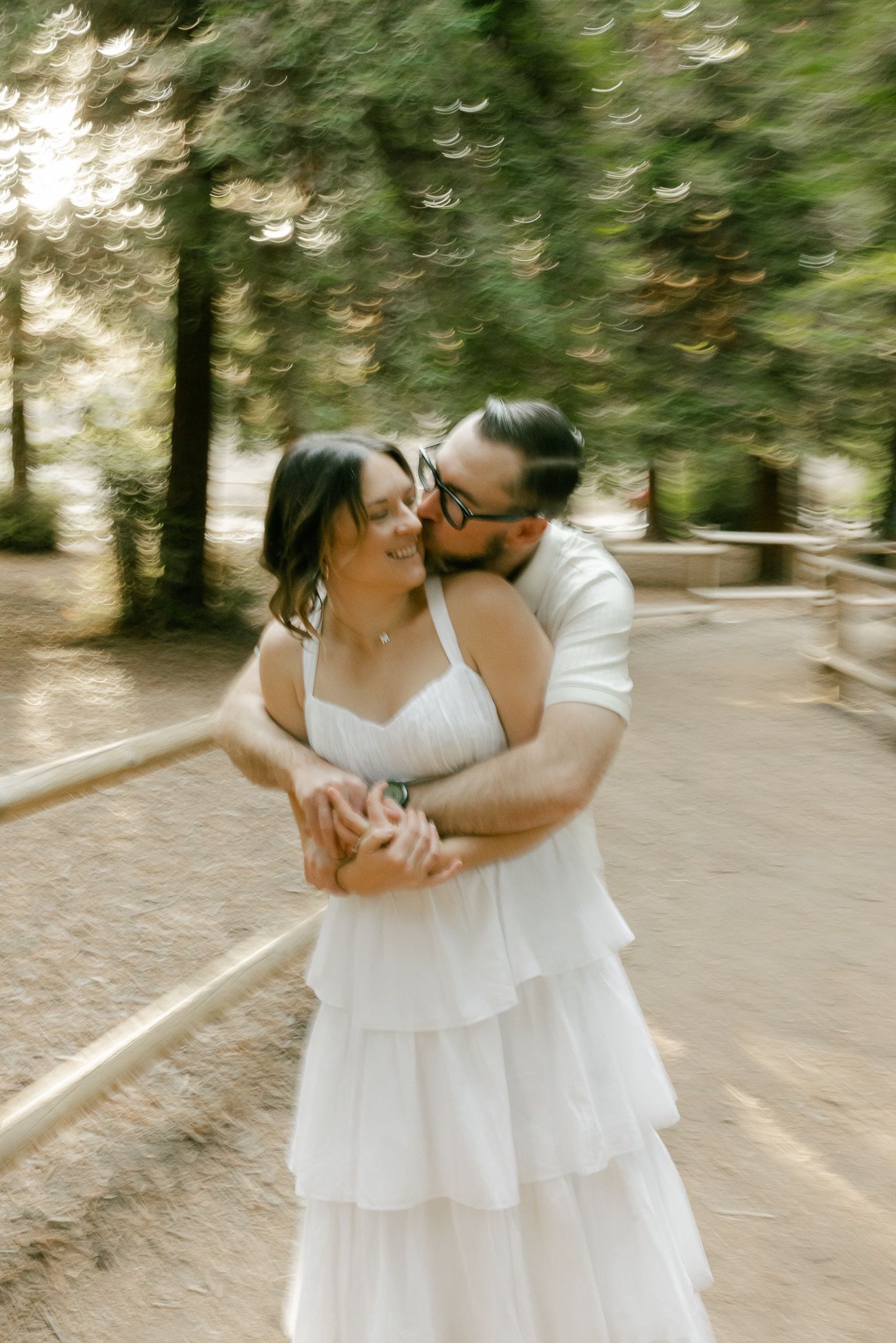 A romantic couple embraces on a forest path, with the person in white wearing a tiered wedding dress.
