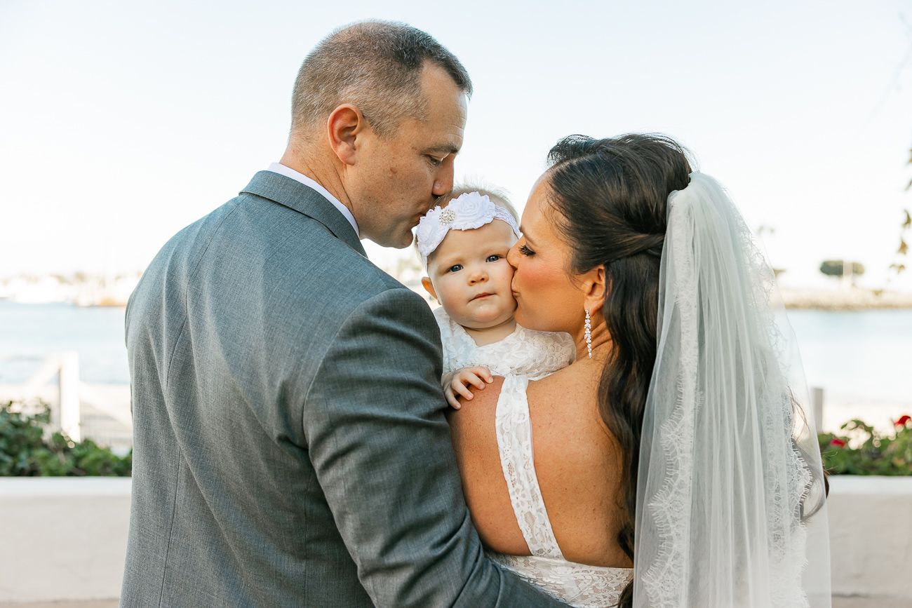 A heartwarming family moment as newlyweds embrace their baby on a sunny balcony overlooking the water.