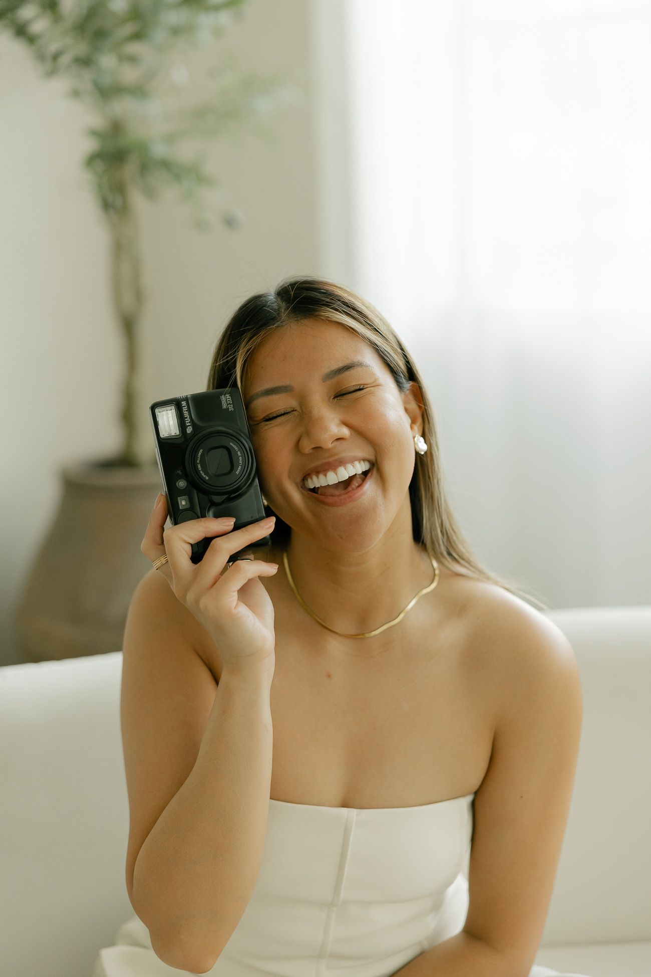 A happy person holding a vintage film camera while sitting on a white couch in natural lighting.