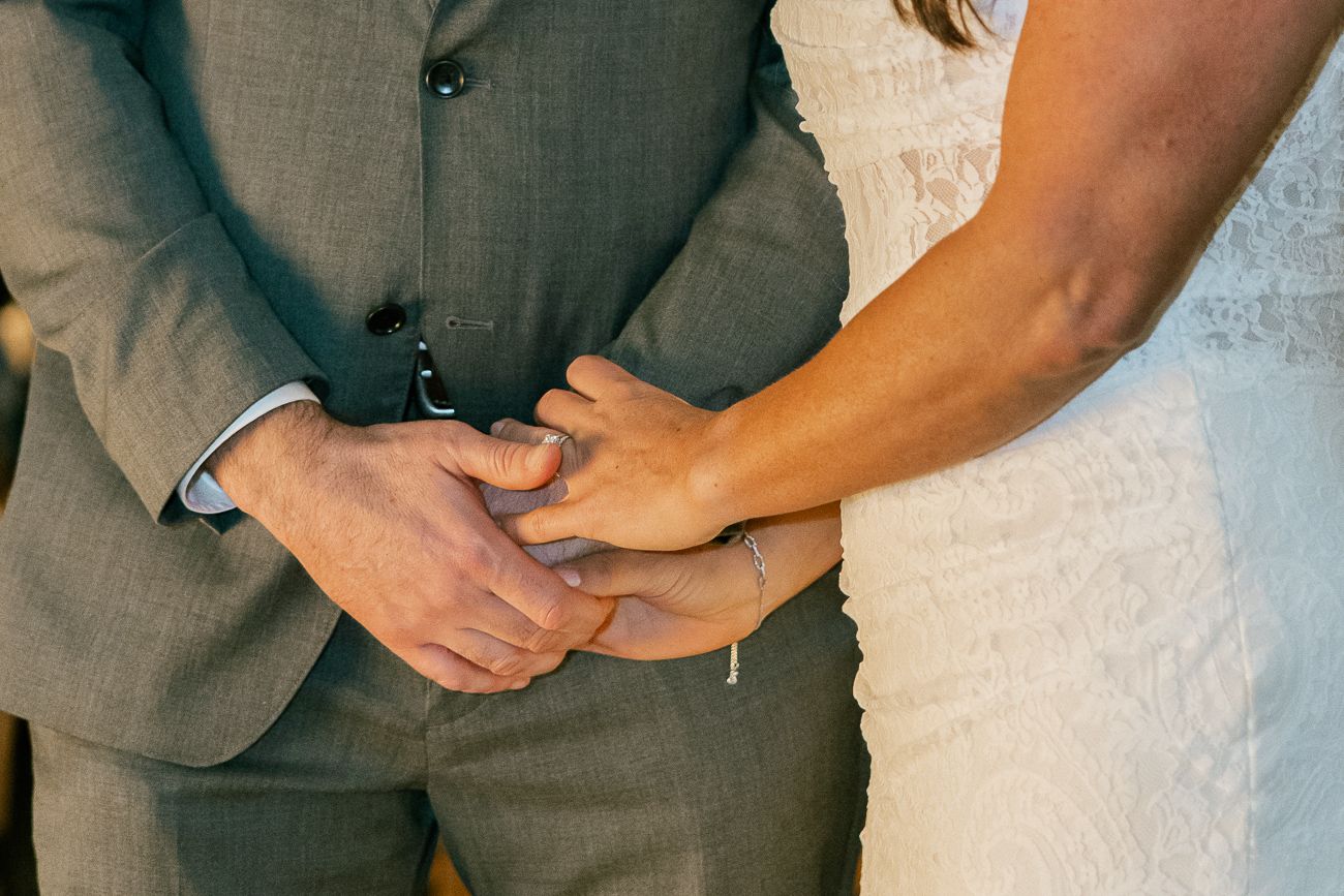 Close-up of hands holding each other, showing a bride in white lace and groom in gray suit.