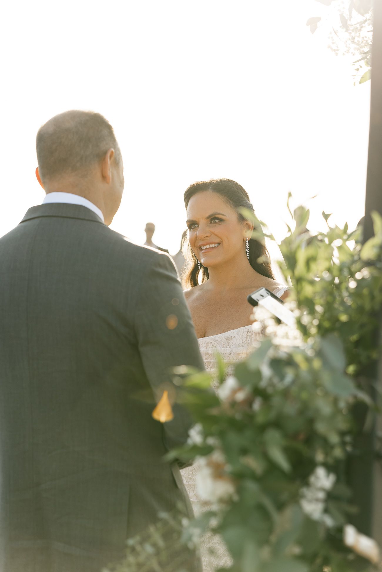 A couple shares a tender moment at an outdoor wedding ceremony, surrounded by soft sunlight and greenery.