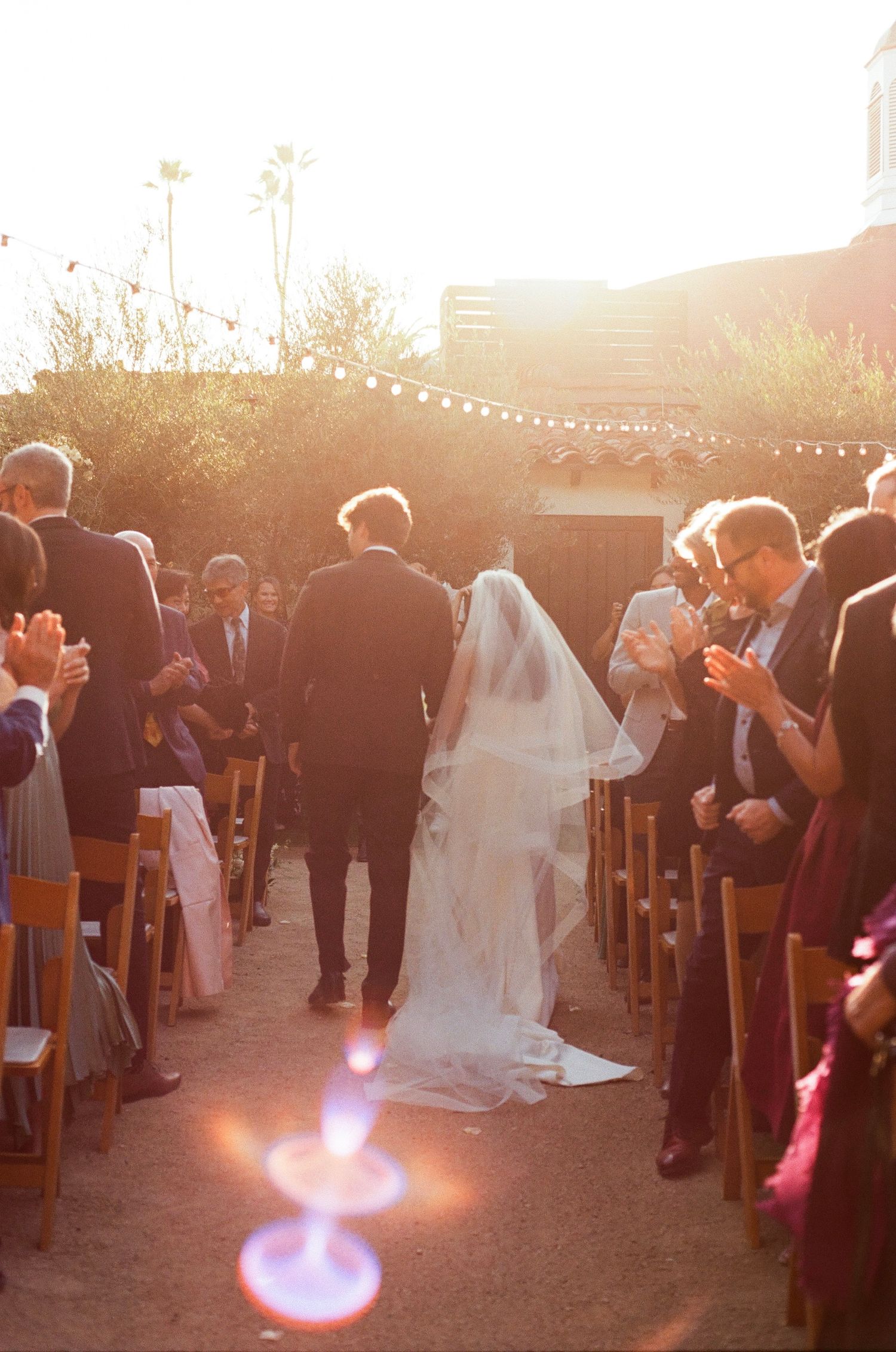 An outdoor wedding ceremony at sunset with string lights overhead and guests standing along wooden chairs.