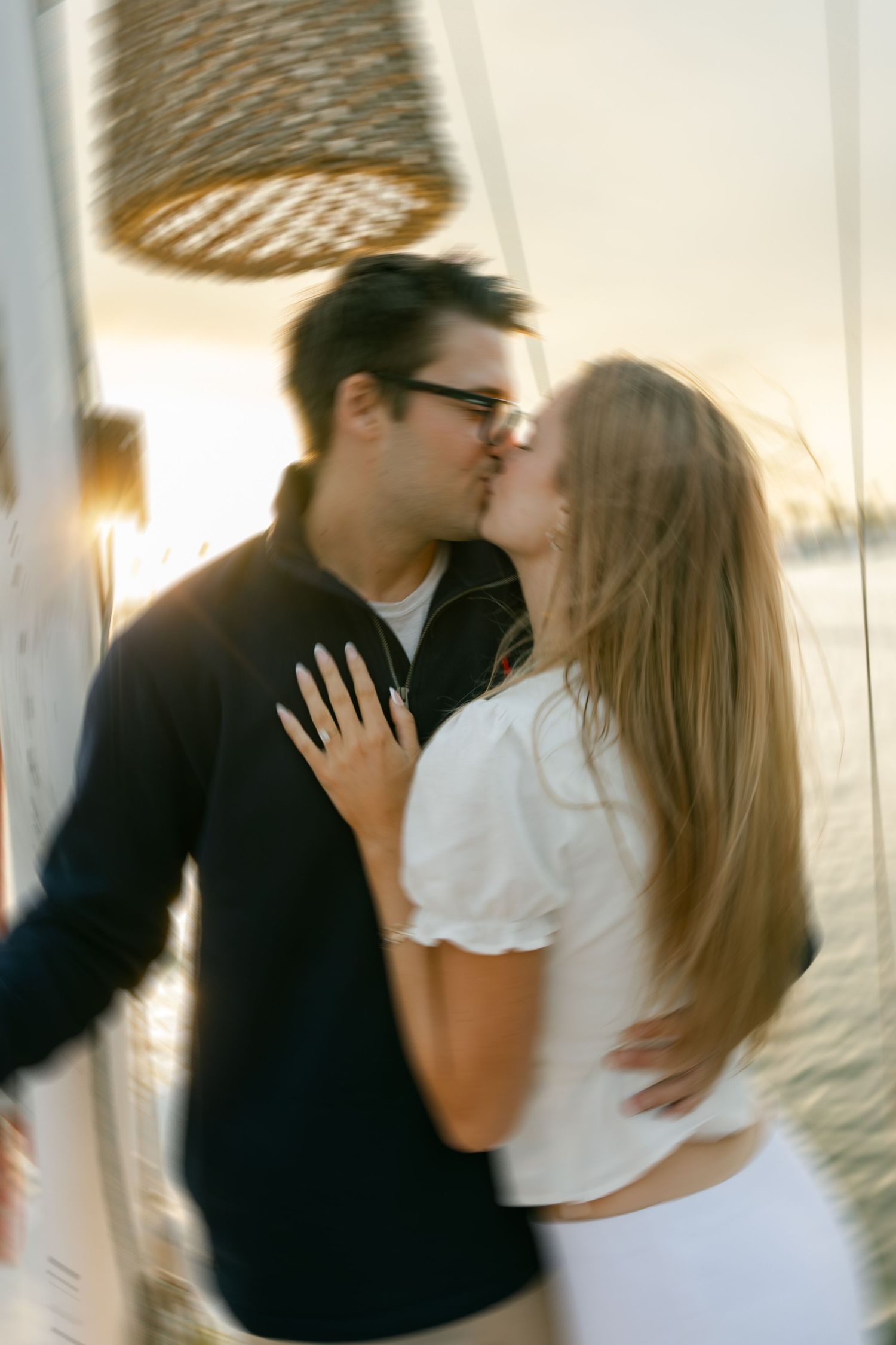 A romantic couple shares a tender kiss at sunset with a woven lamp fixture visible above them.