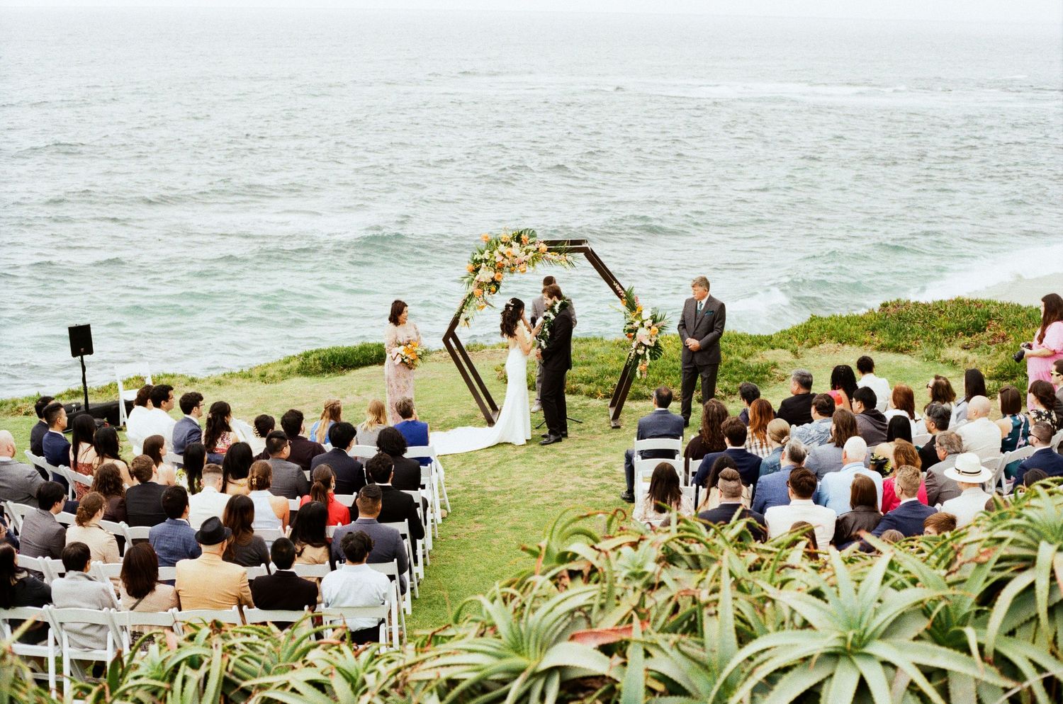 A scenic outdoor wedding ceremony overlooking the ocean with guests seated in white chairs on a grassy cliff.