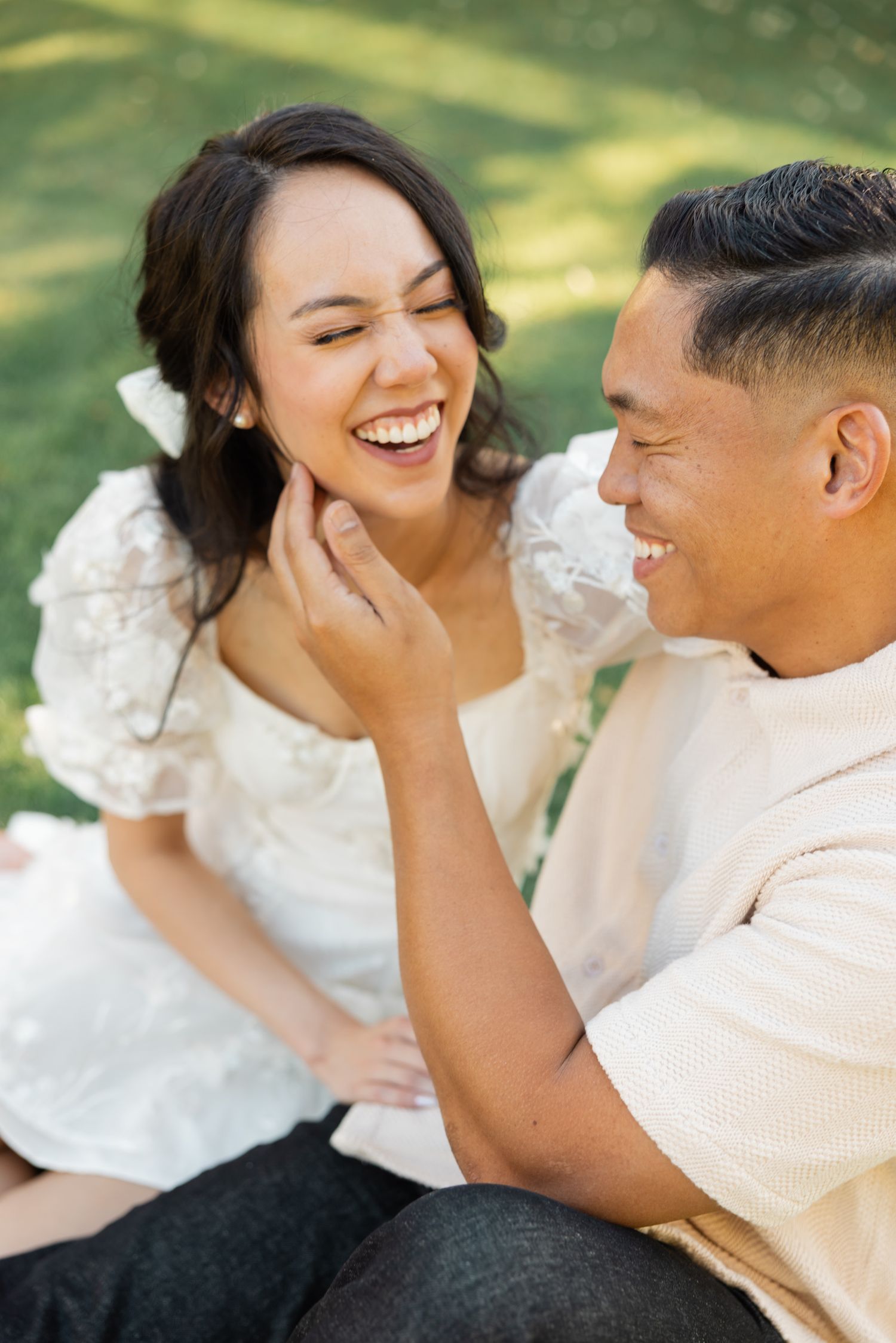 A couple shares intimate laughter while sitting together outdoors in matching white outfits.