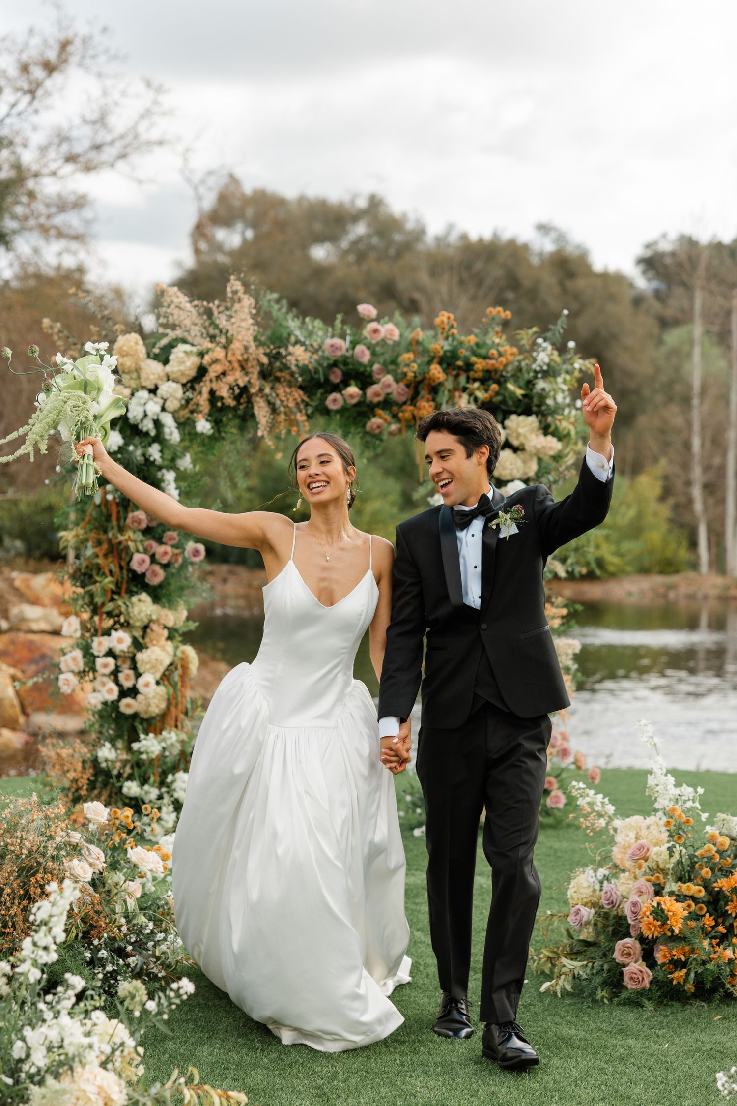Wedding celebration under a floral arch with blooming flowers and greenery by a scenic pond.