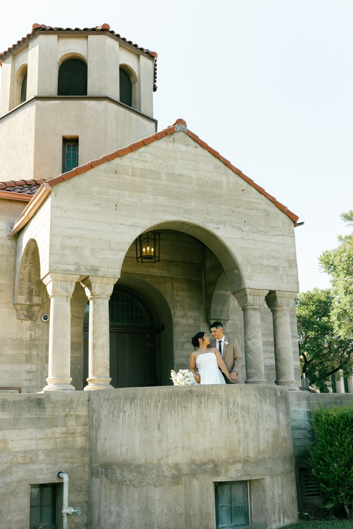 Spanish style church with white stucco walls and terracotta roof features a couple on its arched portico.