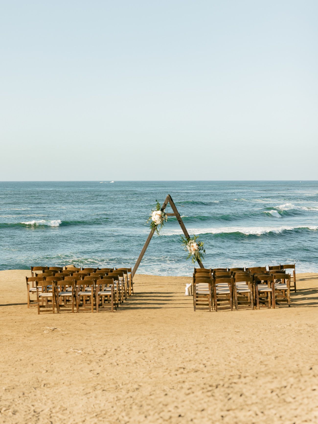 Wooden chairs set up for a beach wedding ceremony with a triangular arch and ocean waves in the background.