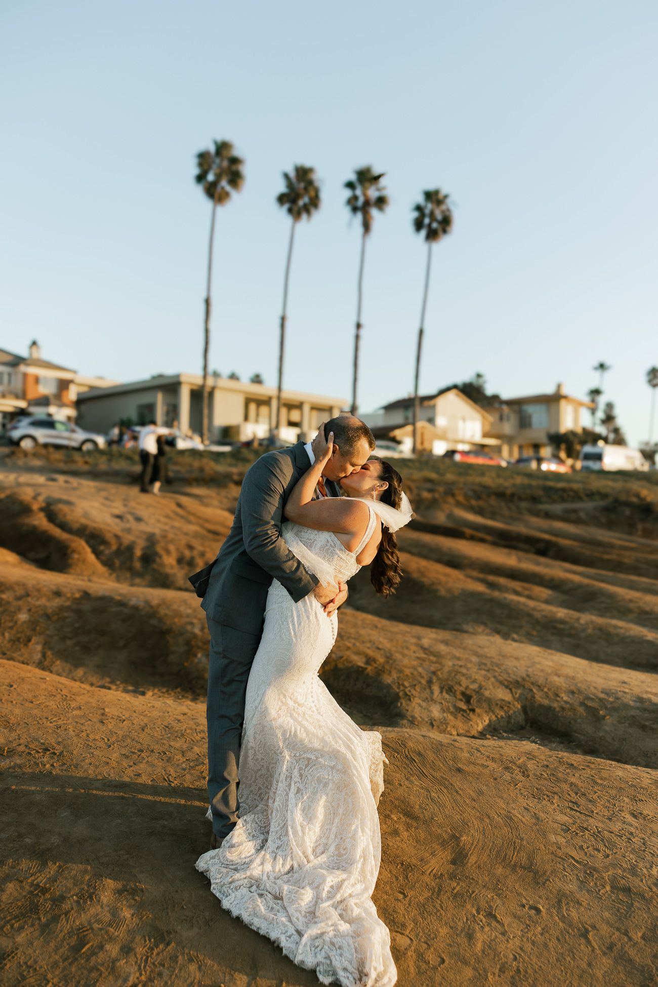 Newlyweds share an intimate embrace on a beach cliff with palm trees and coastal homes in the background at sunset.