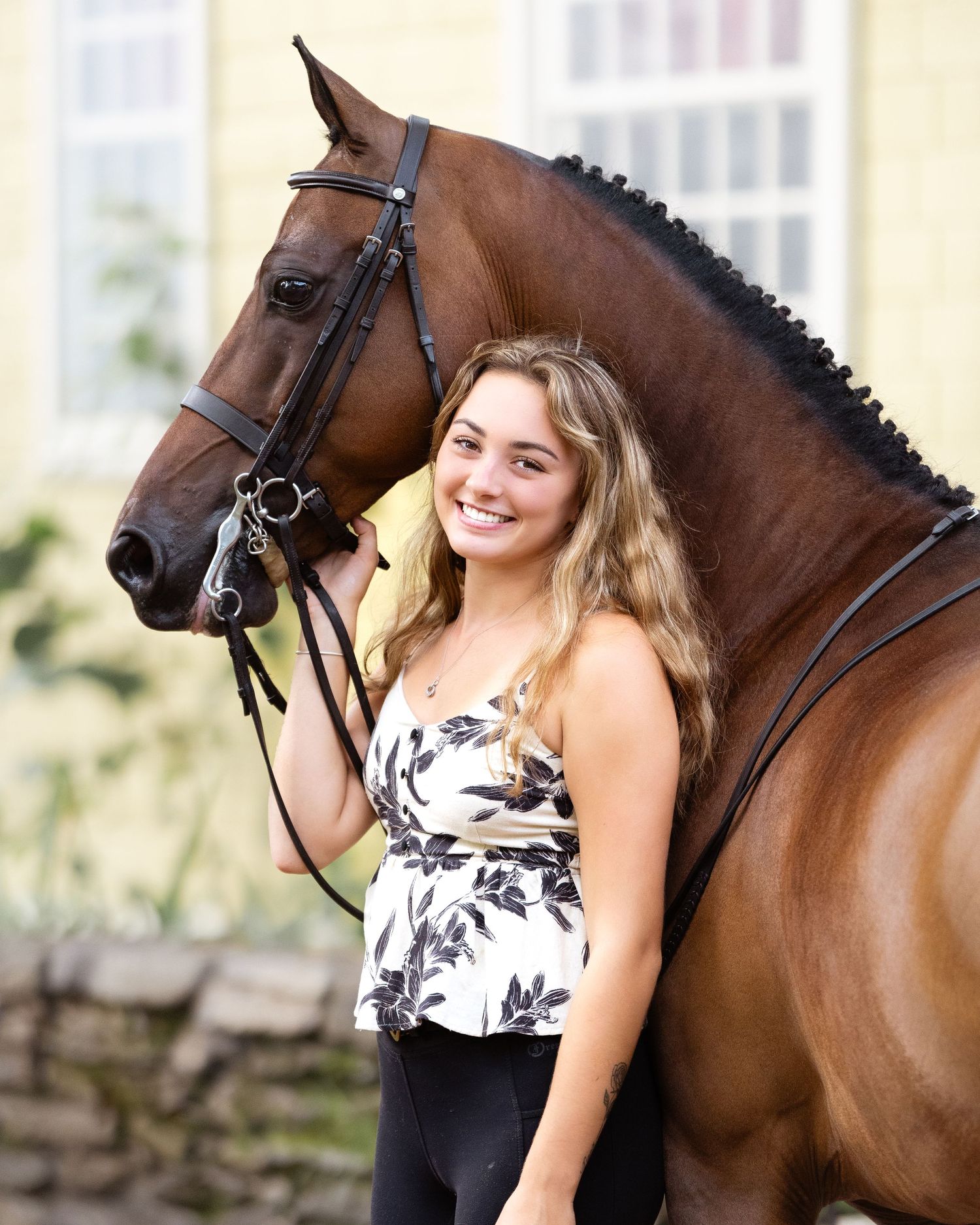 A majestic bay horse with braided mane stands beside its owner for a portrait against a stone wall outdoors.