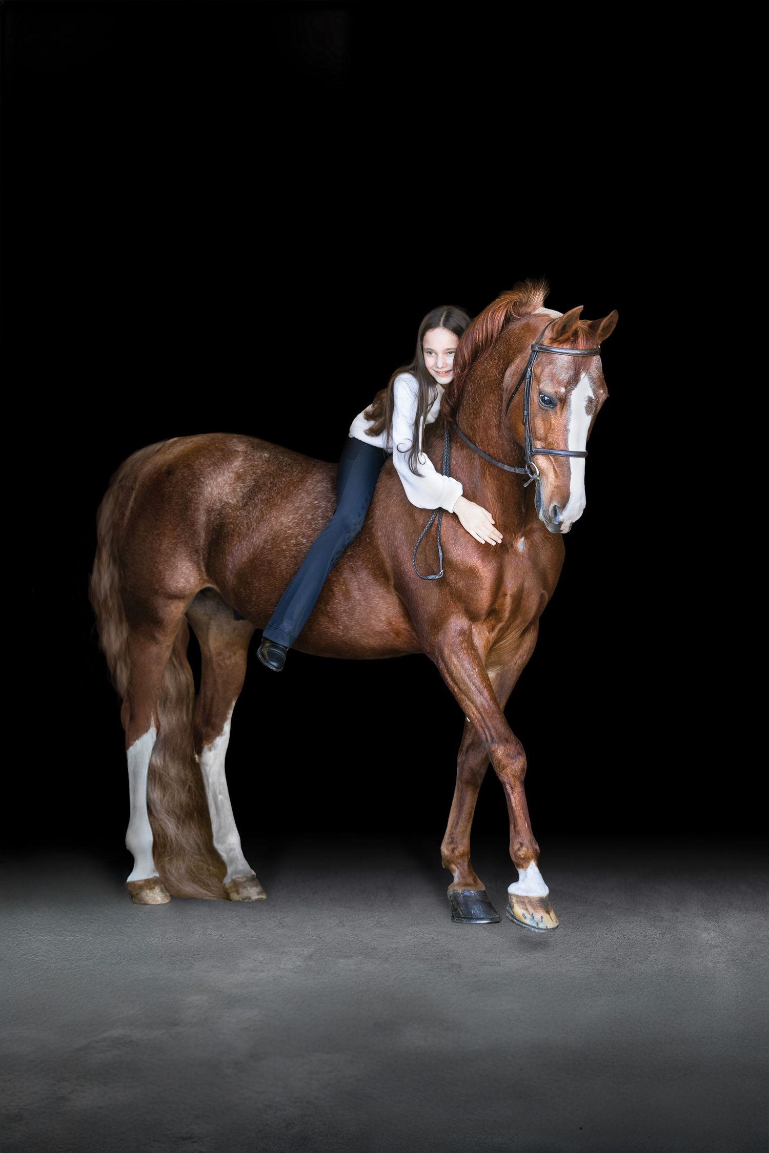 A young girl in equestrian attire hugs a chestnut horse while riding bareback against a dark studio background.