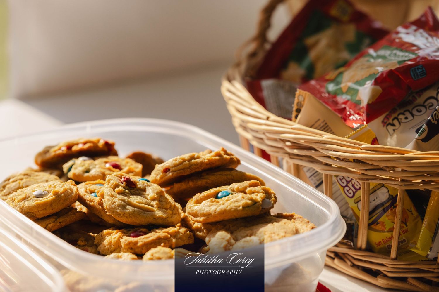 Fresh baked chocolate chip cookies in a plastic container next to a decorative basket on a counter.
