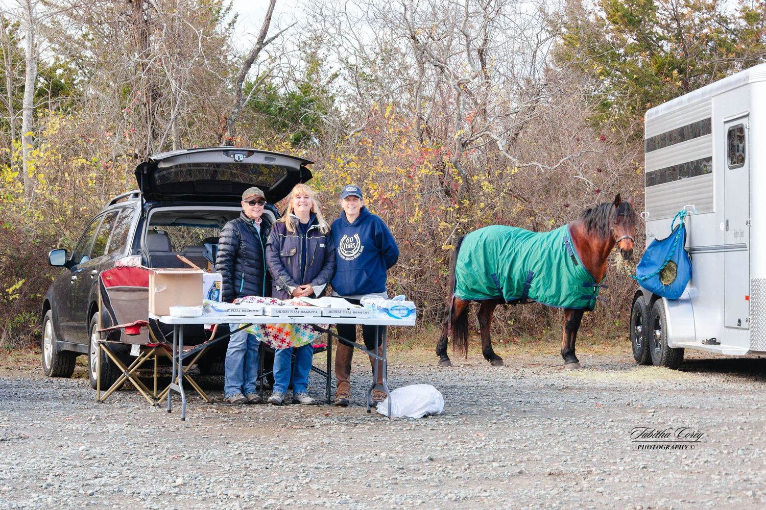 People gathered at a table outside a horse trailer while a blanketed horse stands nearby on a fall day.
