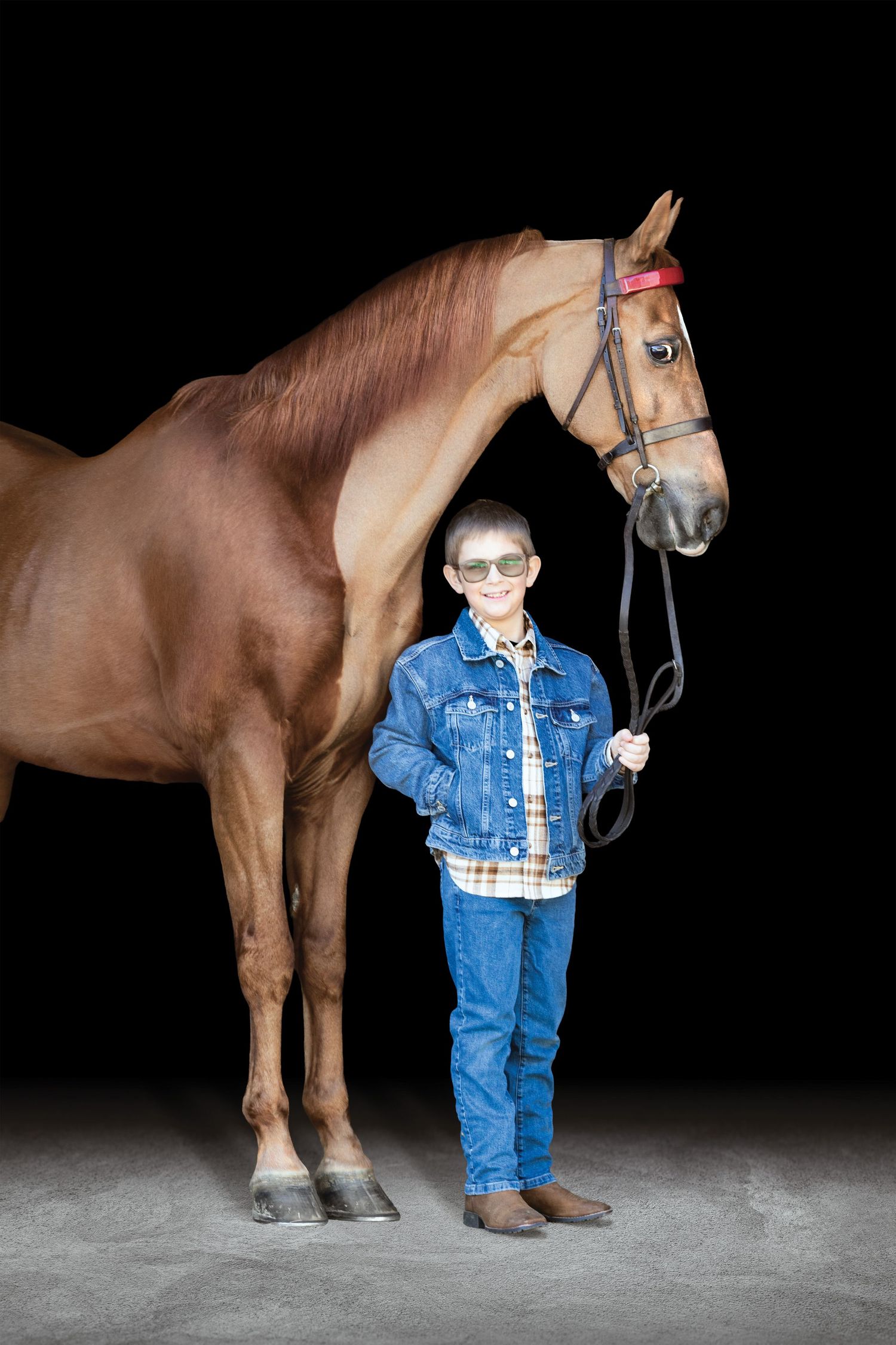A smiling boy in denim jacket and jeans stands beside a chestnut horse against a black background.