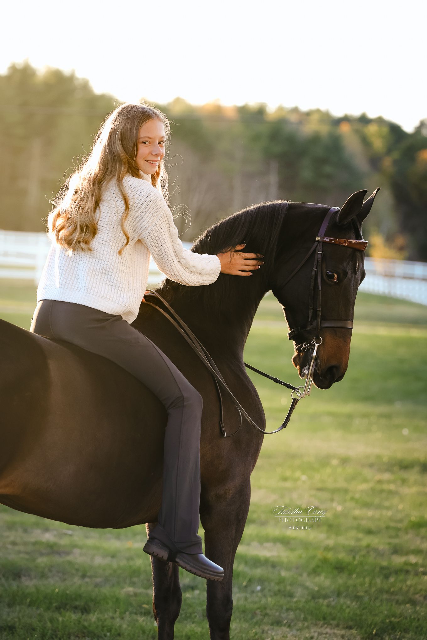 Young girl in white sweater riding a dark bay horse at golden hour in a green pasture with white fence.