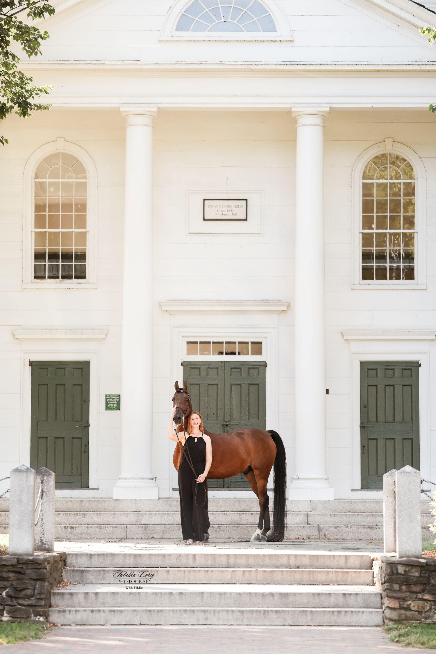 Woman in black outfit stands with a brown horse on steps of a white colonial building with green doors.