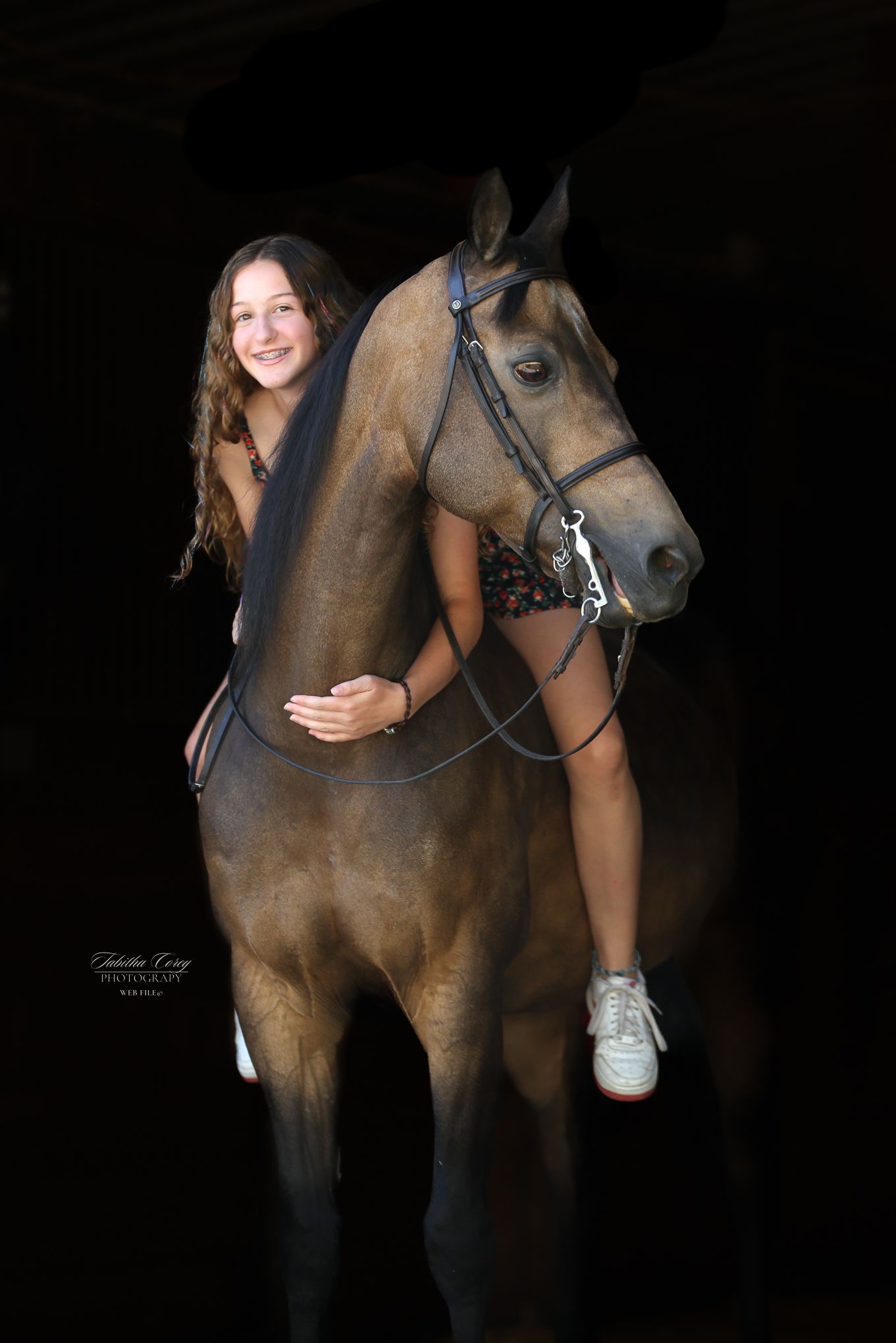 A rider photographed against a black background sits atop a majestic brown horse wearing a black leather bridle.
