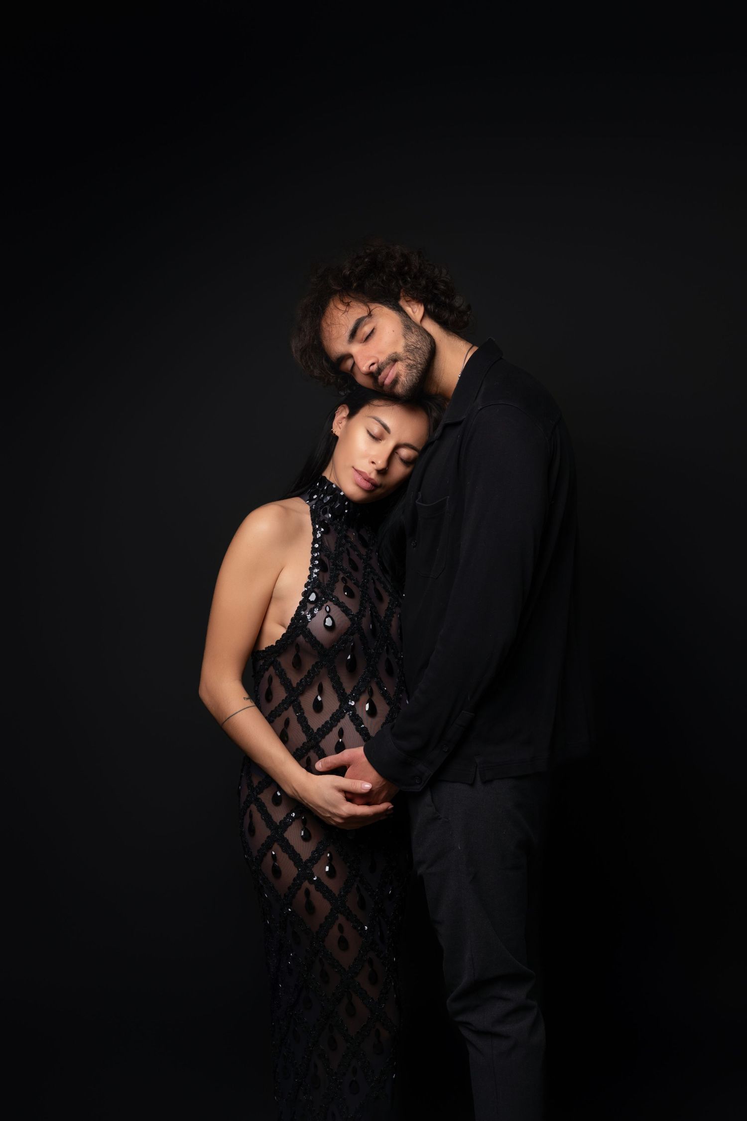 A couple embraces intimately against a dark background, with the woman wearing a patterned dress and man in dark clothing.