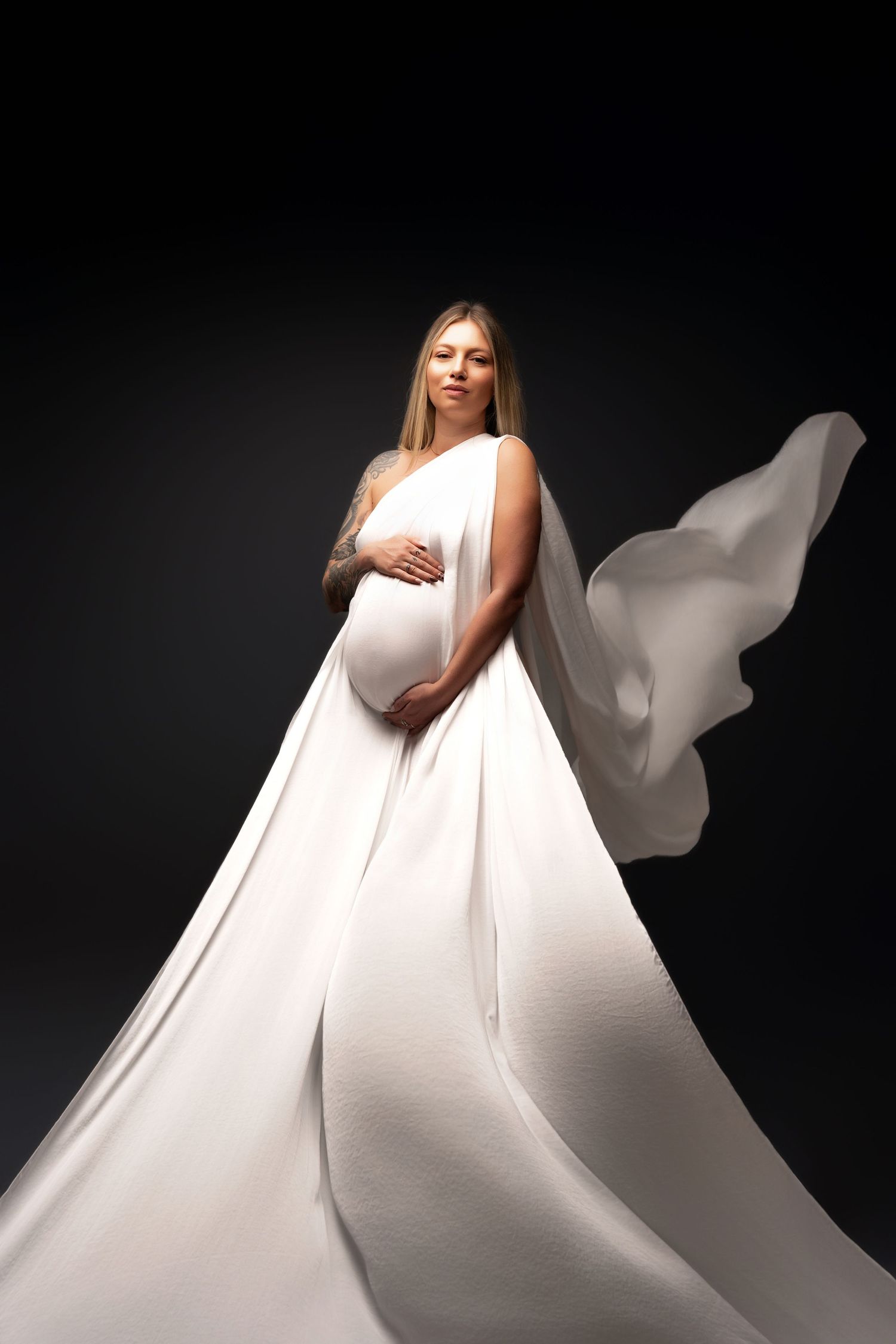 A pregnant figure in a flowing white gown poses elegantly against a dark background as fabric billows dramatically.