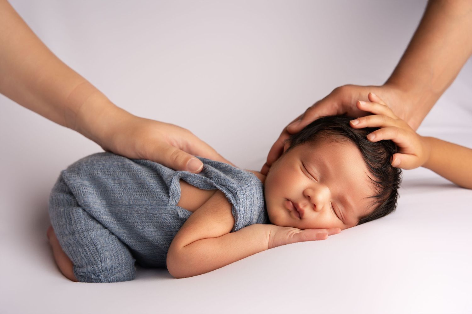A sleeping newborn baby in gray romper lays on white background while hands gently touch head and back.