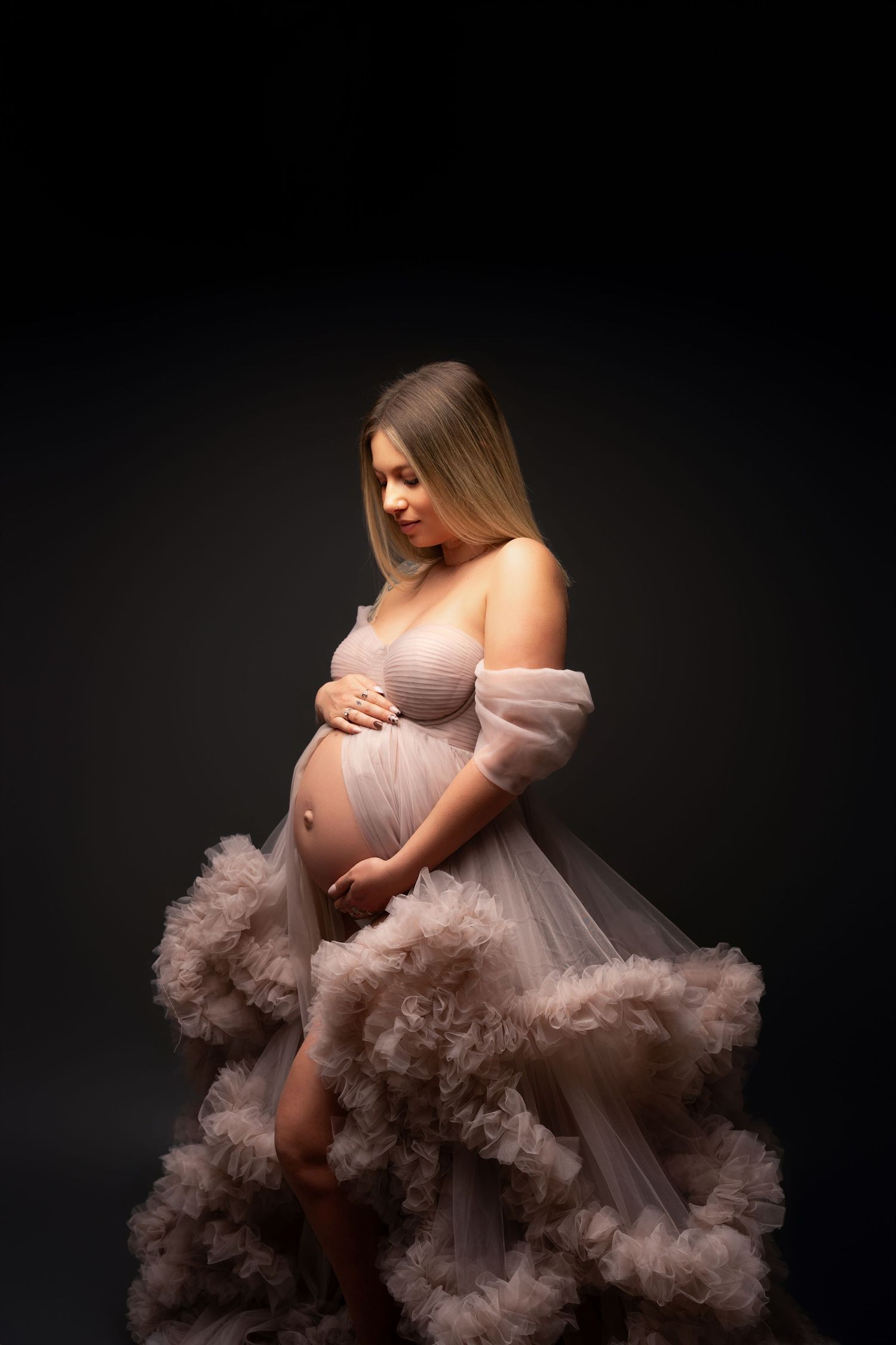 A model in a flowing blush pink tulle gown poses against a dark backdrop in a dramatic fashion shoot.