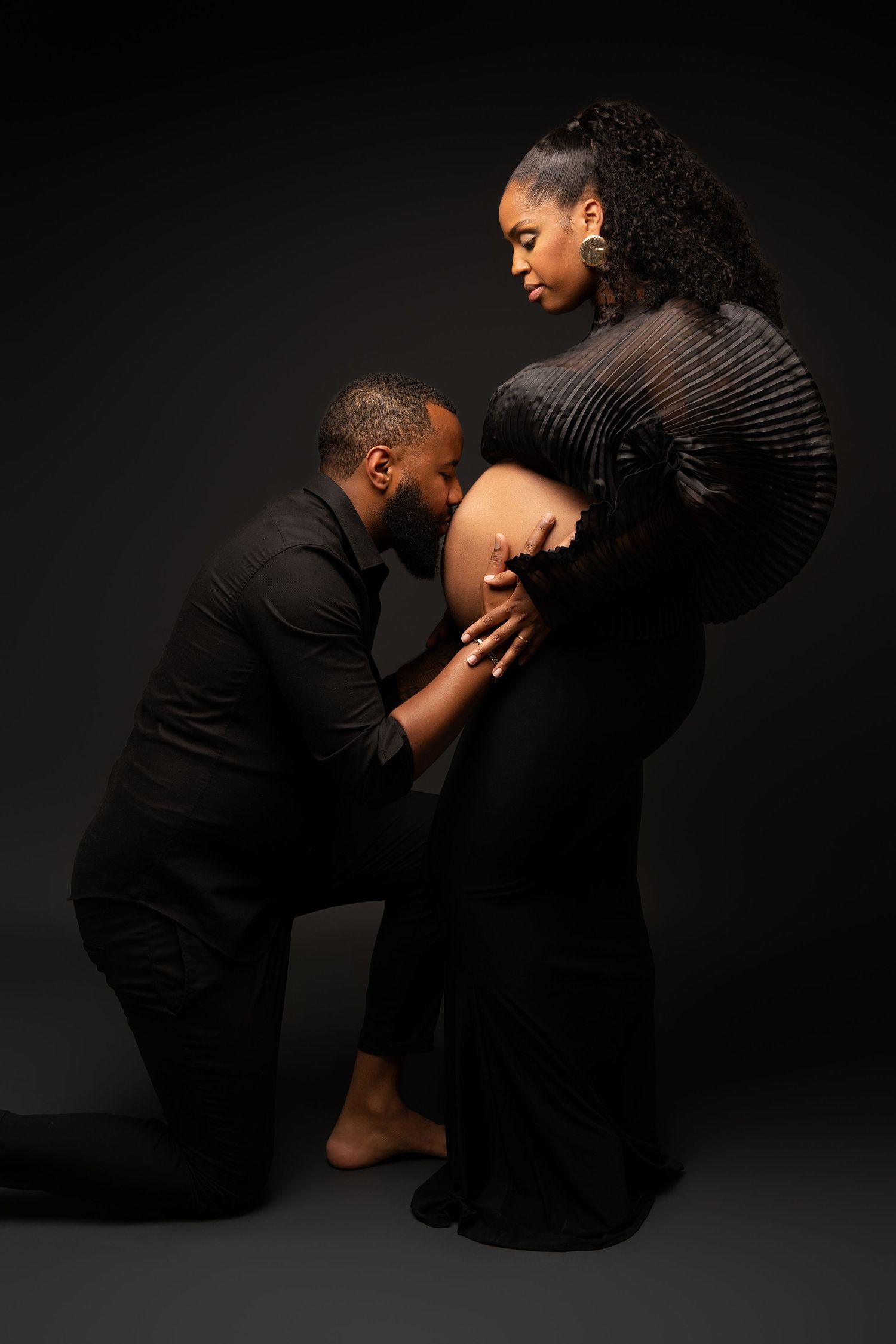 Intimate maternity photo shoot with couple wearing black against dark background.