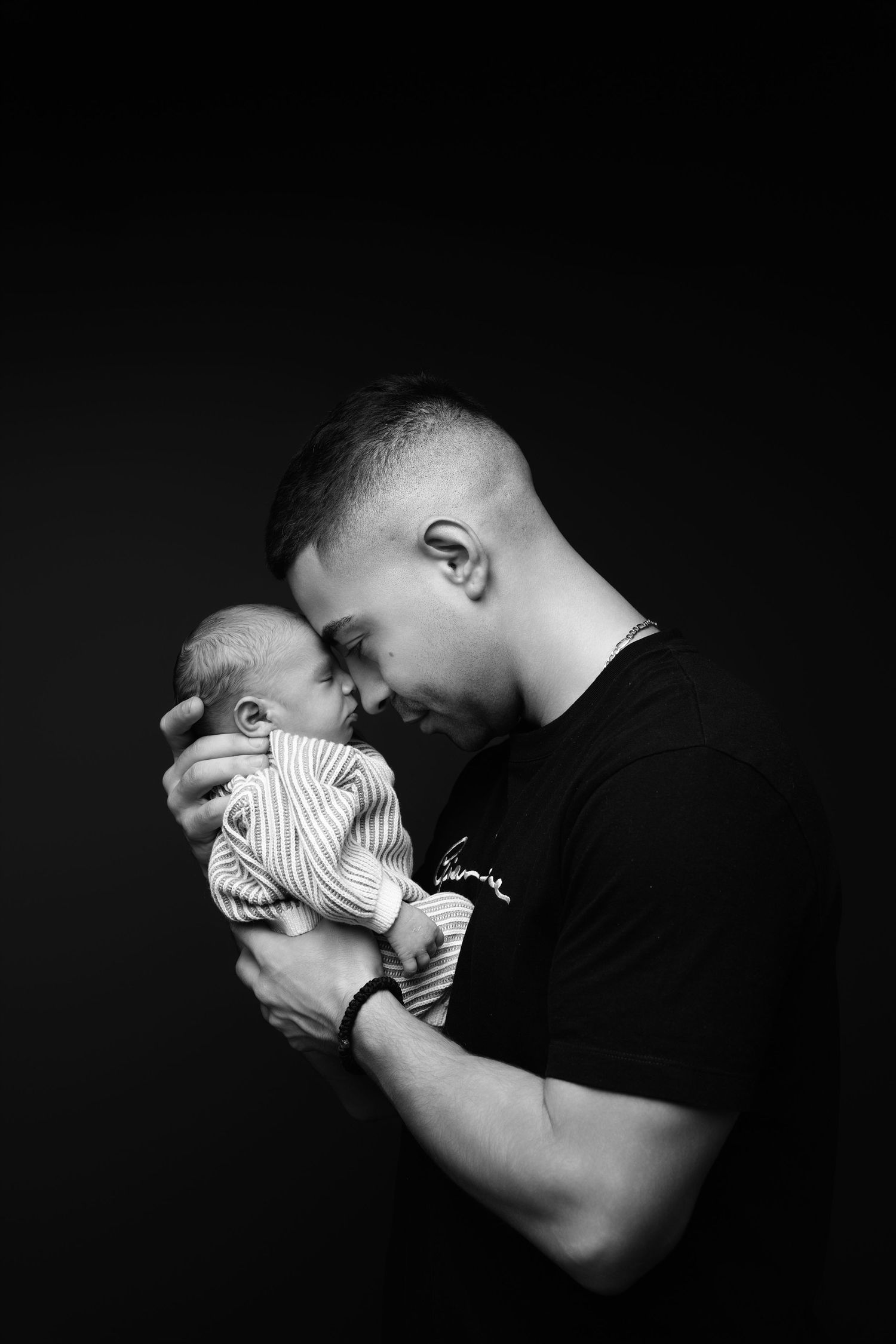 Black and white artistic photo series showing tender moments between parent and newborn in dark dramatic lighting.
